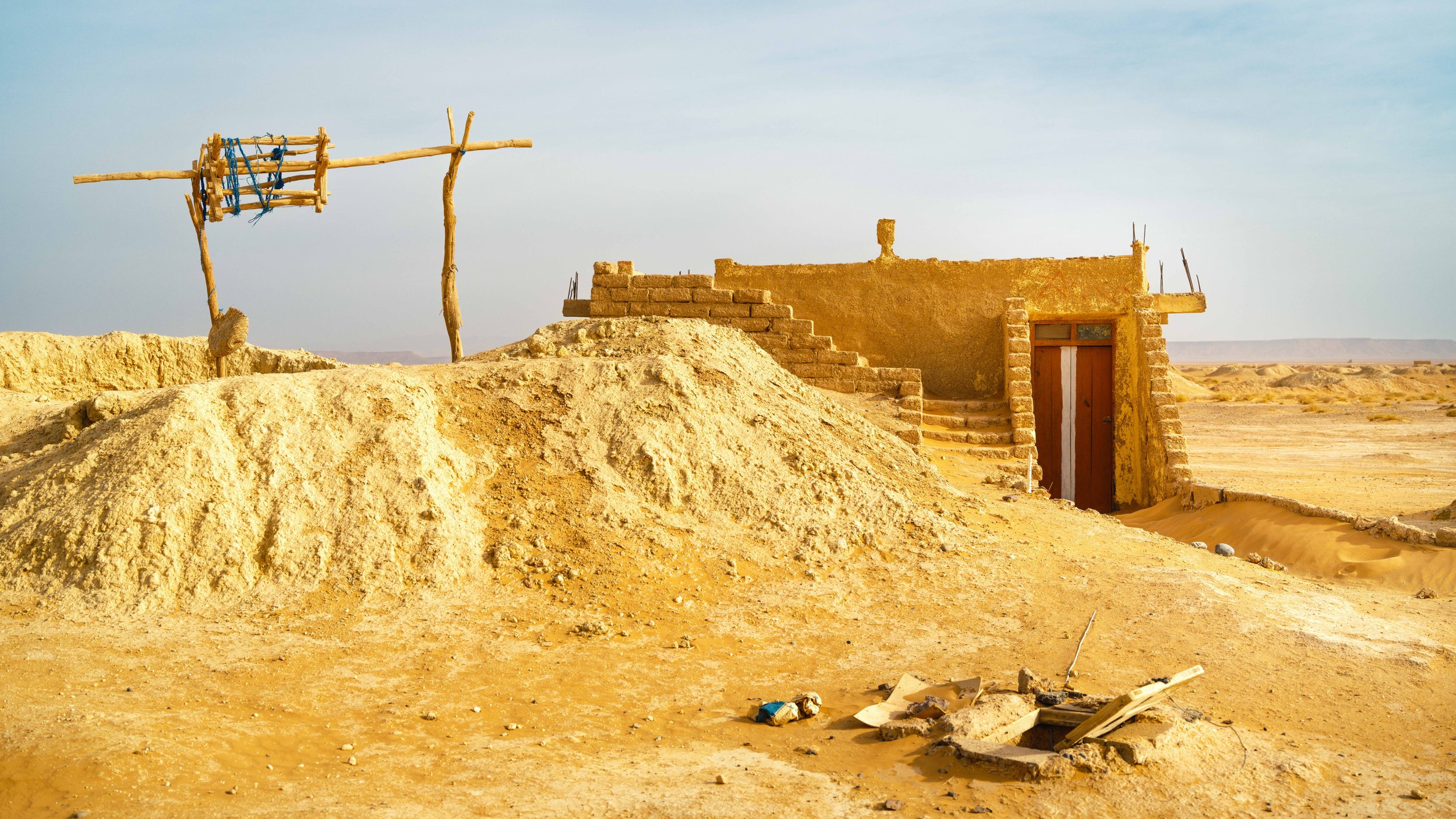 A small, worn adobe house with a wooden door, surrounded by sand and desert landscape. There are steps leading to the roof and a simple, makeshift wooden antenna structure nearby.
