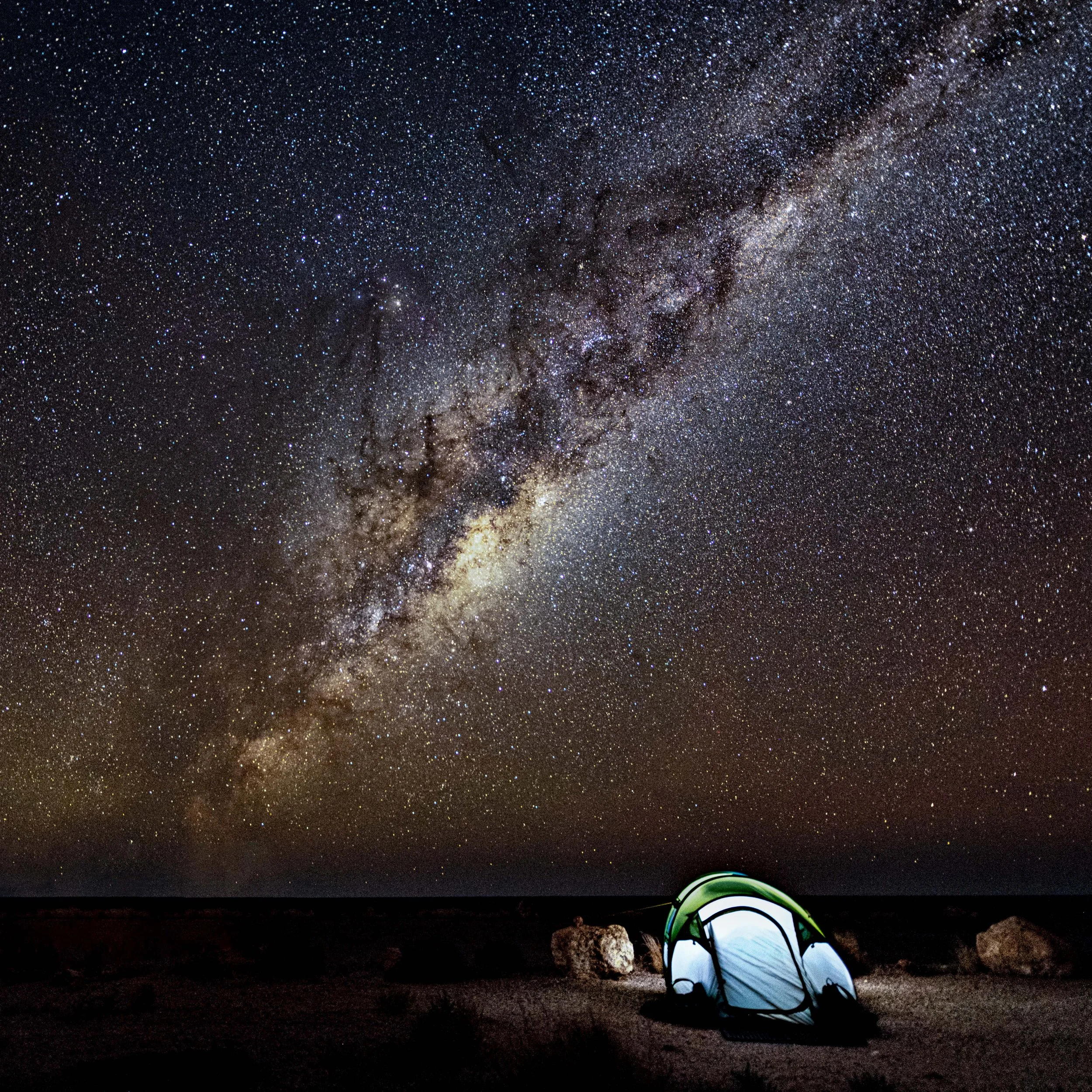 A green and white tent set up on a dark landscape with the Milky Way galaxy visible in the night sky above.
