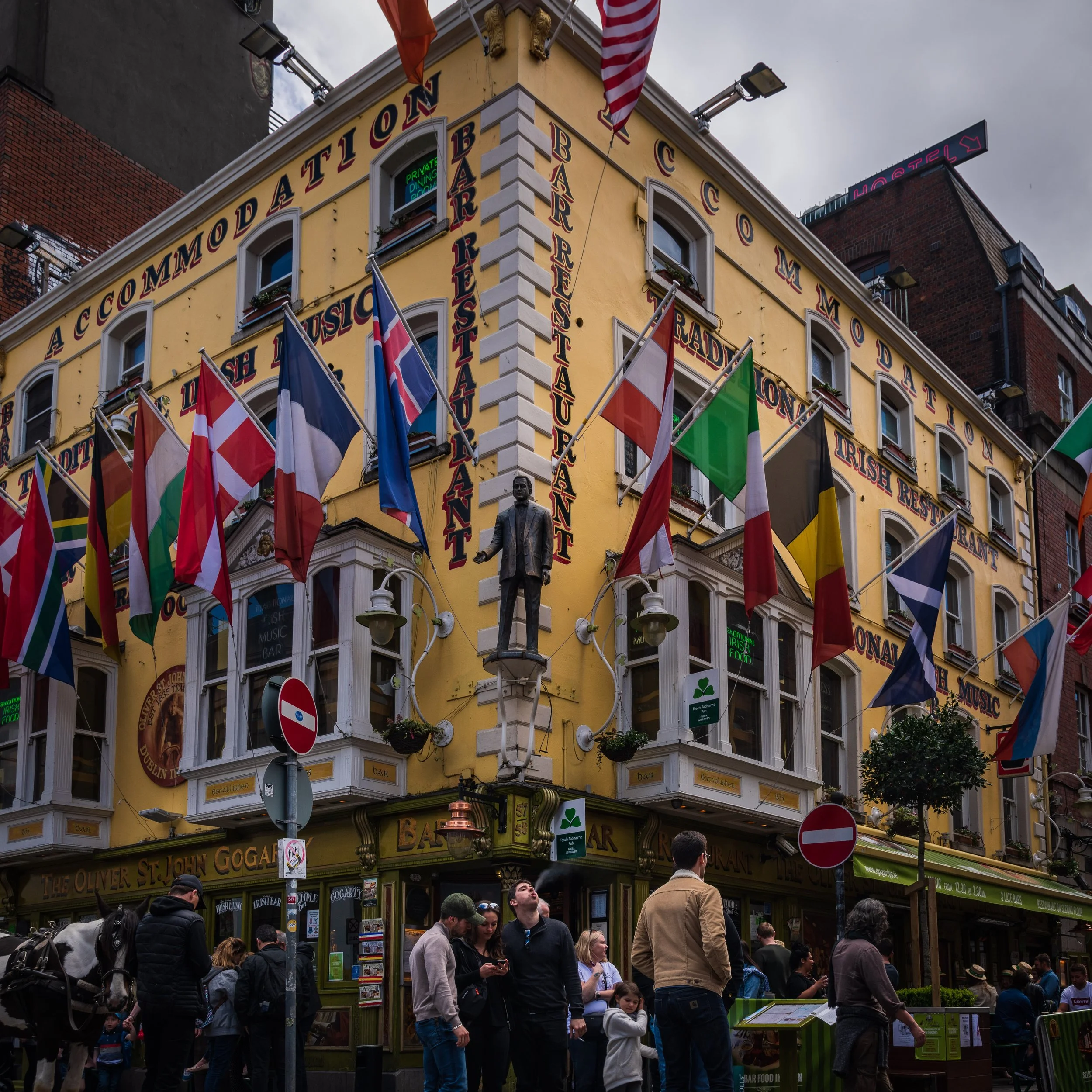 Exterior of a lively Irish pub with a yellow facade, adorned with numerous Irish flags, street signs, and a statue of a man on a pedestal. People are gathered outside the pub on a busy street.