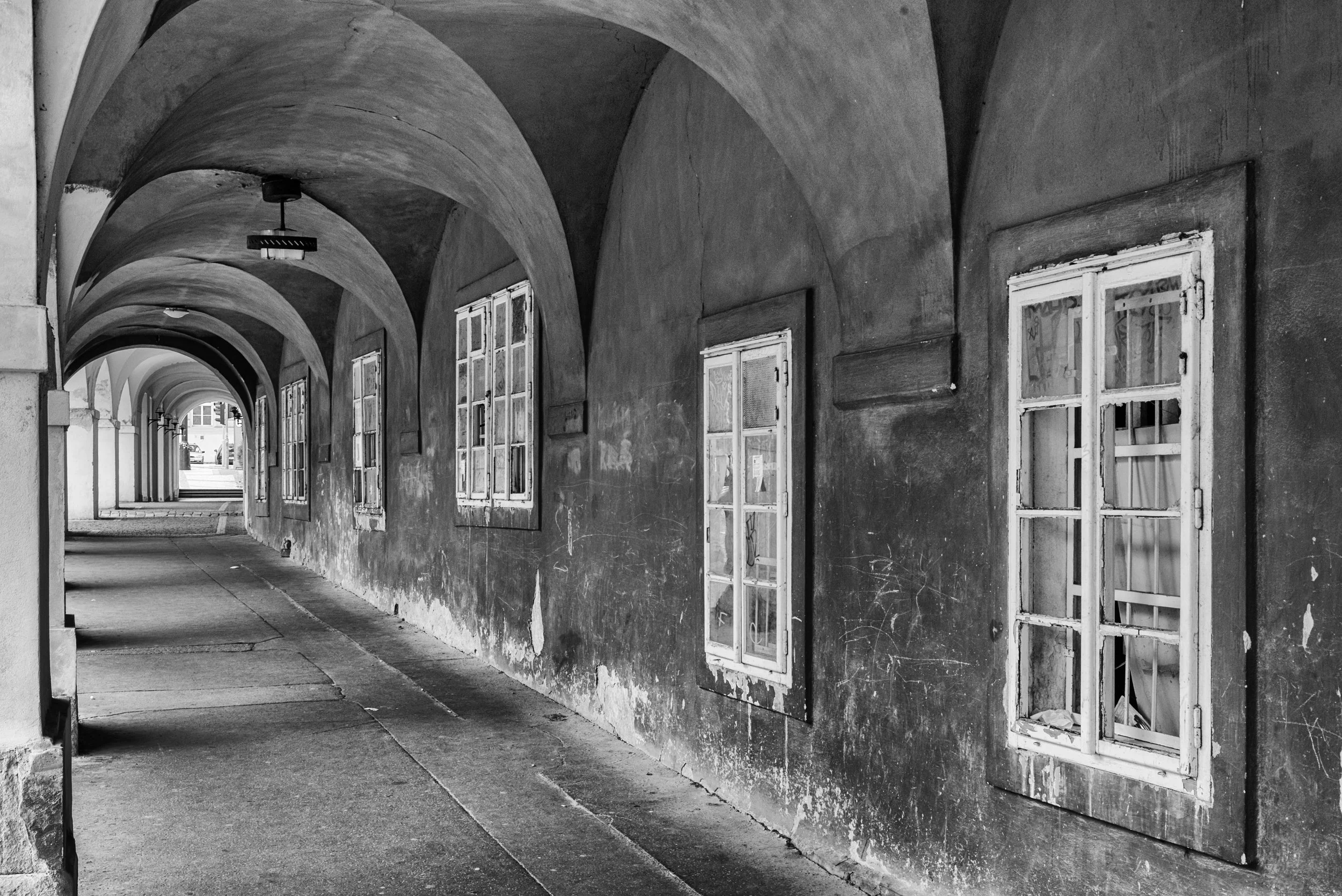Black and white photo of an arched outdoor walkway with multiple worn, open windows along the wall.