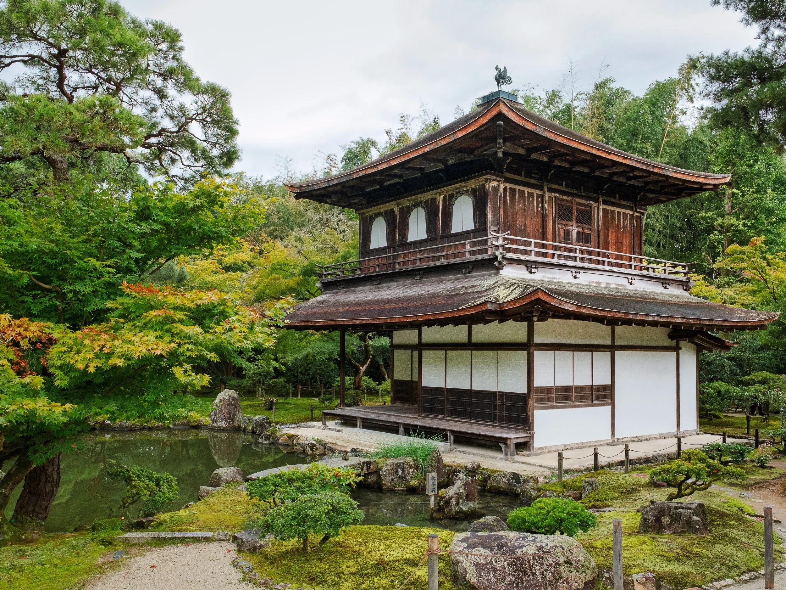 A traditional Japanese wooden building surrounded by lush greenery and a small pond in a garden.
