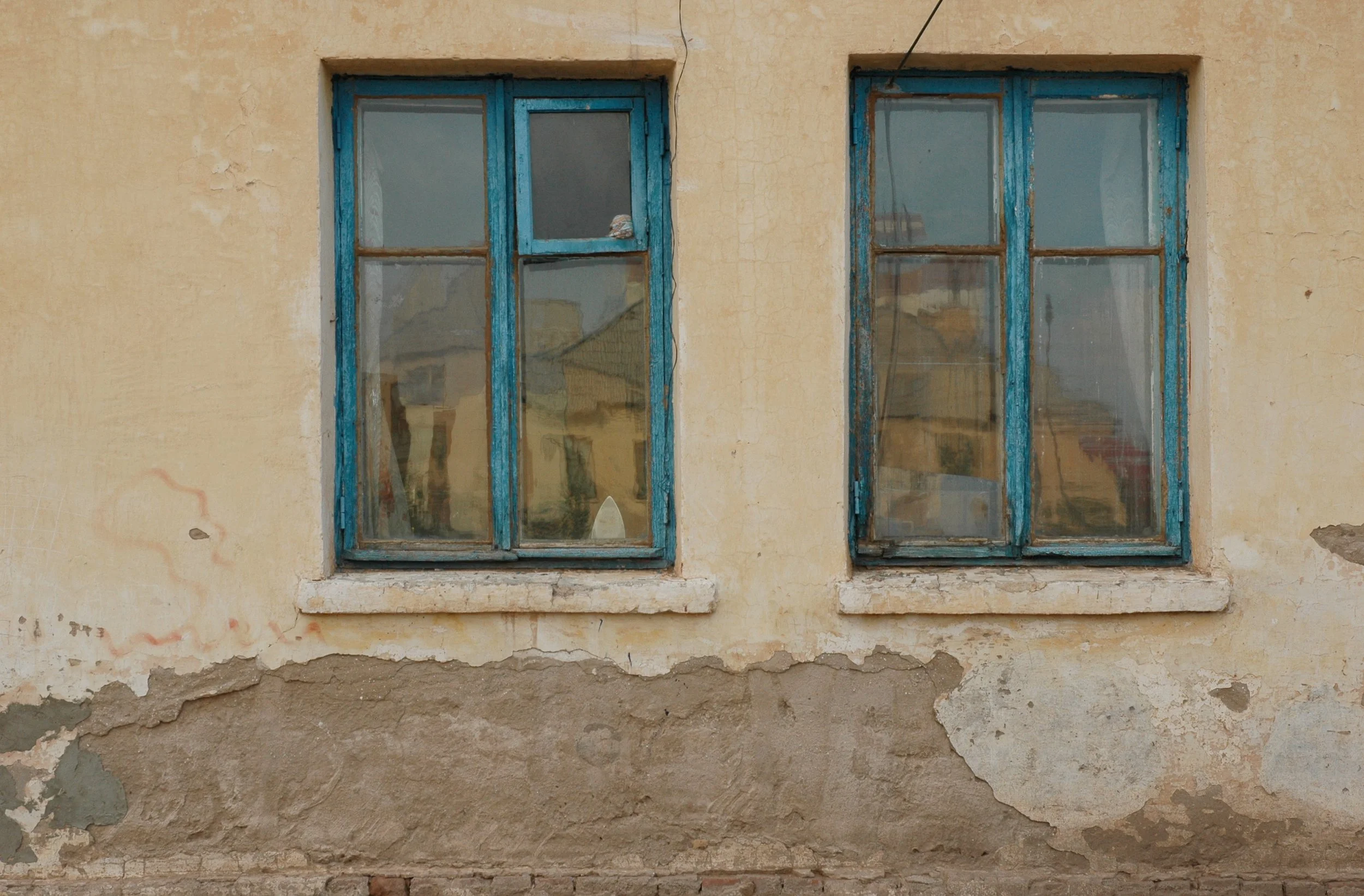 Two old, weathered windows with blue frames on a beige, peeling wall of a building.