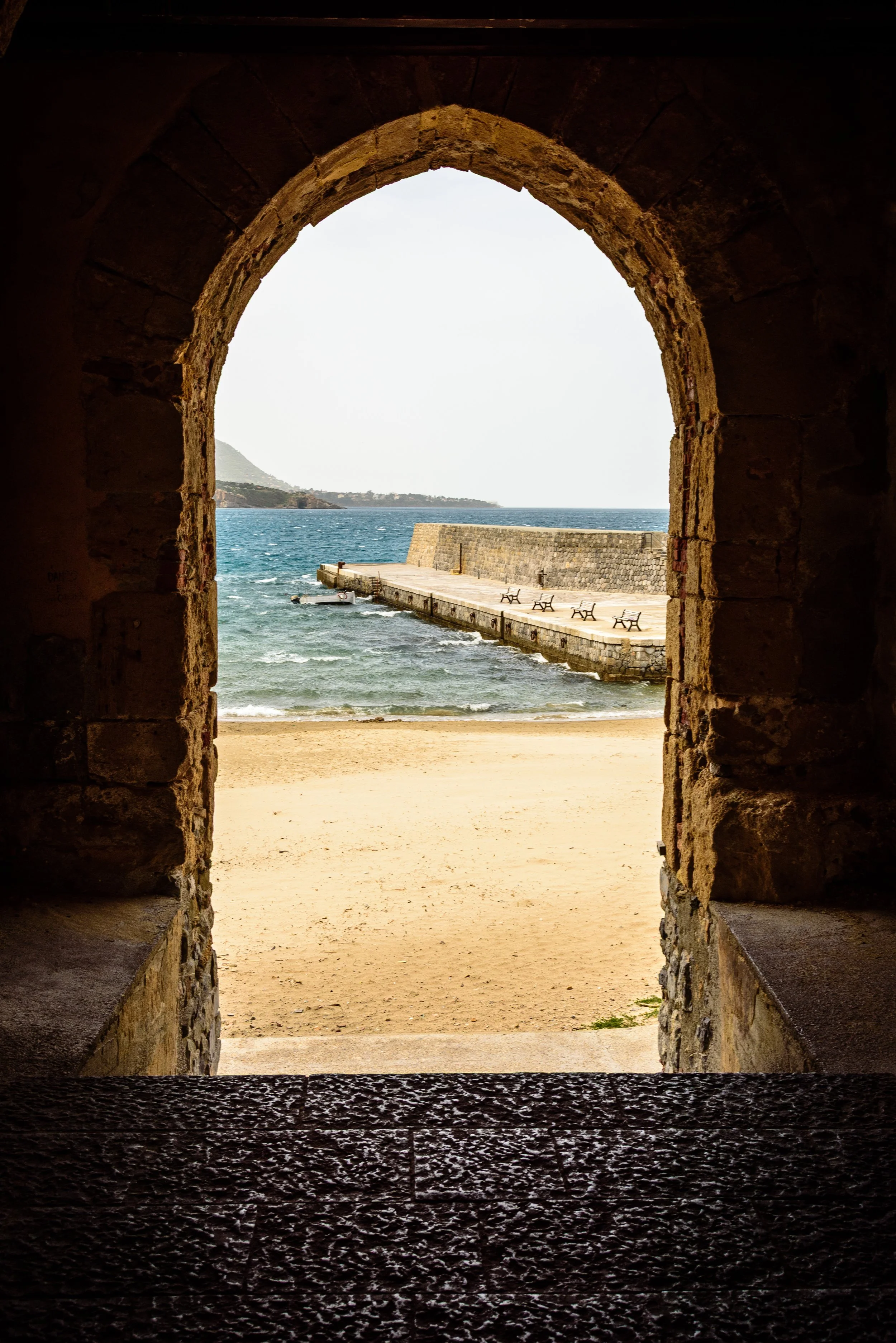 View through a stone archway looking out onto a sandy beach, ocean, and a pier with benches.