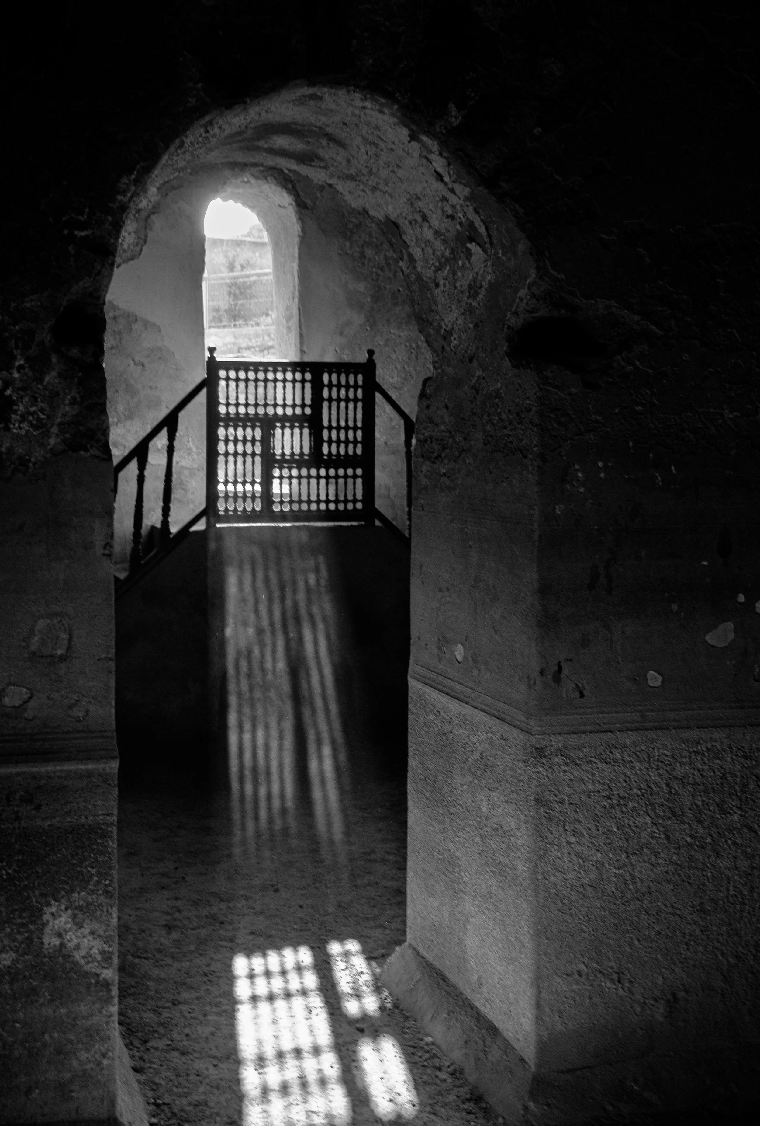 Black and white photograph of an arched doorway in a stone cave, with sunlight creating shadows through a decorative wooden gate and window at the top of a staircase.