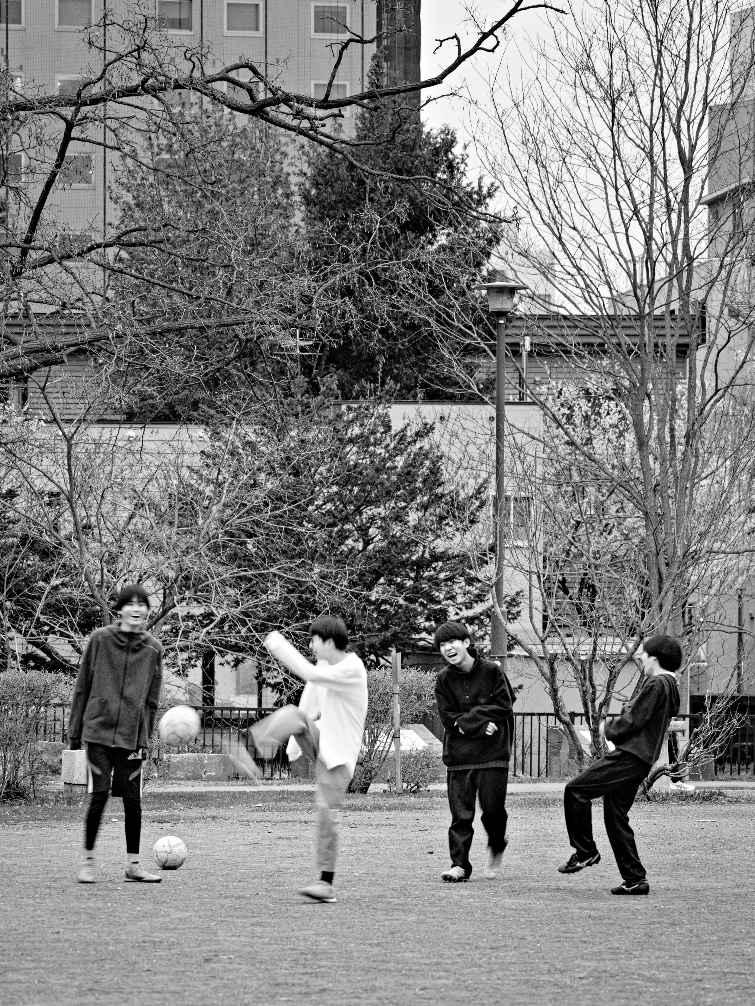 Four boys playing soccer outdoors on a grassy field with leafless trees and buildings in the background.