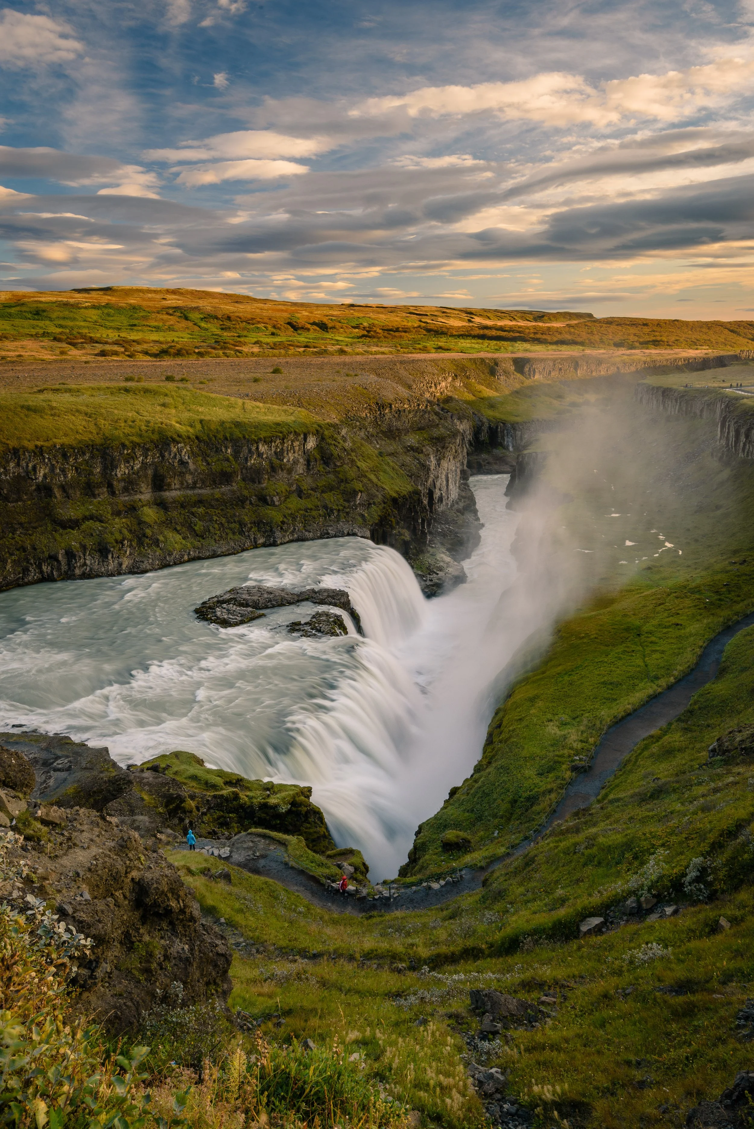 Gullfoss waterfall in Iceland with cascading water, mist, green landscape, and cloudy sky.