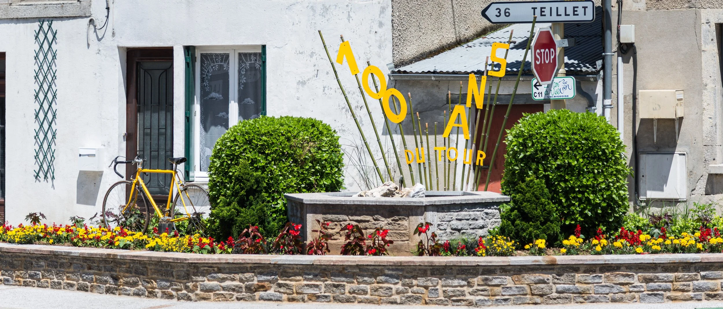 A decorated outdoor planter with green bushes, red and yellow flowers, and yellow street art spelling '100 N' in front of a white building with bicycle and street signs.
