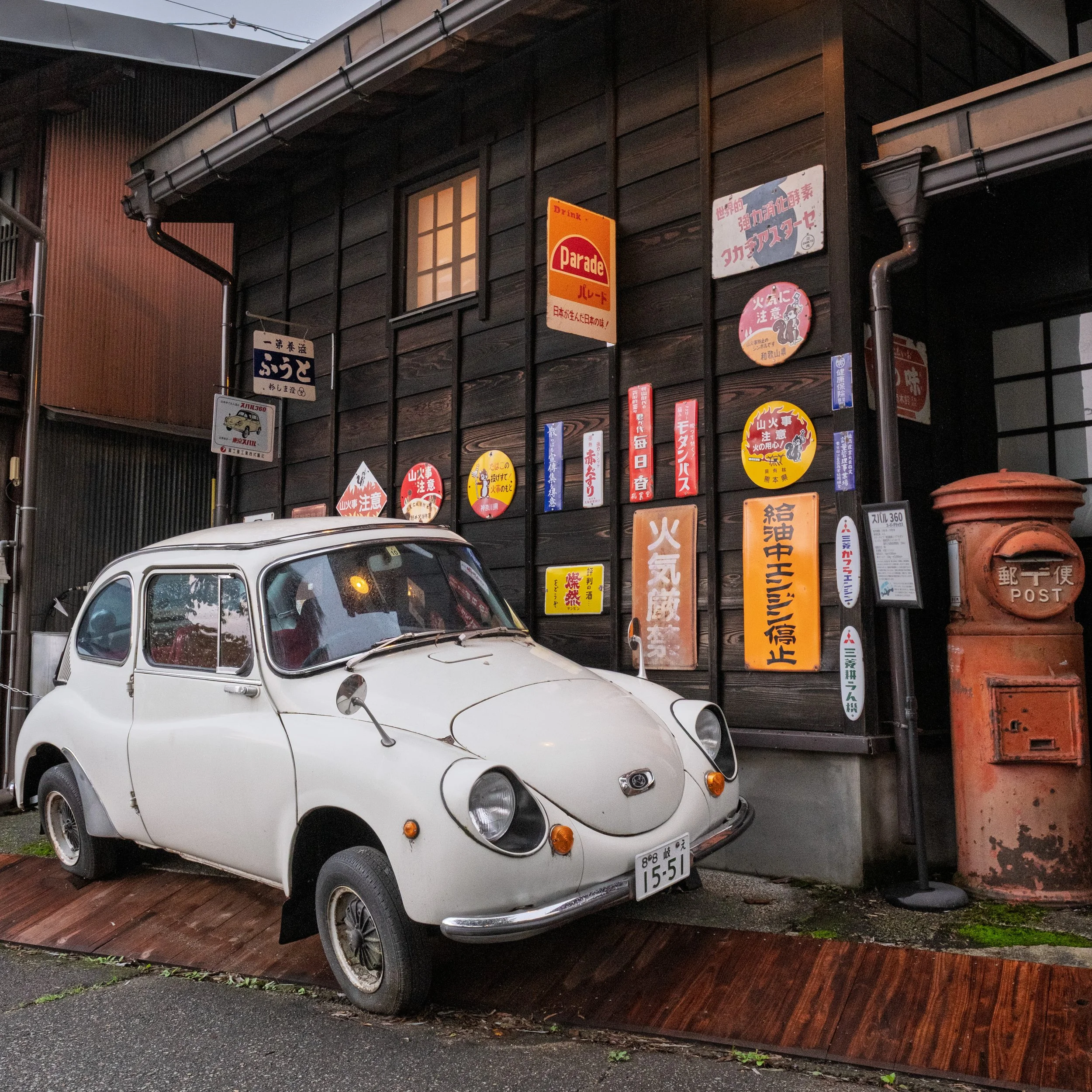 A vintage white car parked on a wooden ramp outside a dark wooden building with Japanese signs and posters on the wall. An orange post box is beside the car.