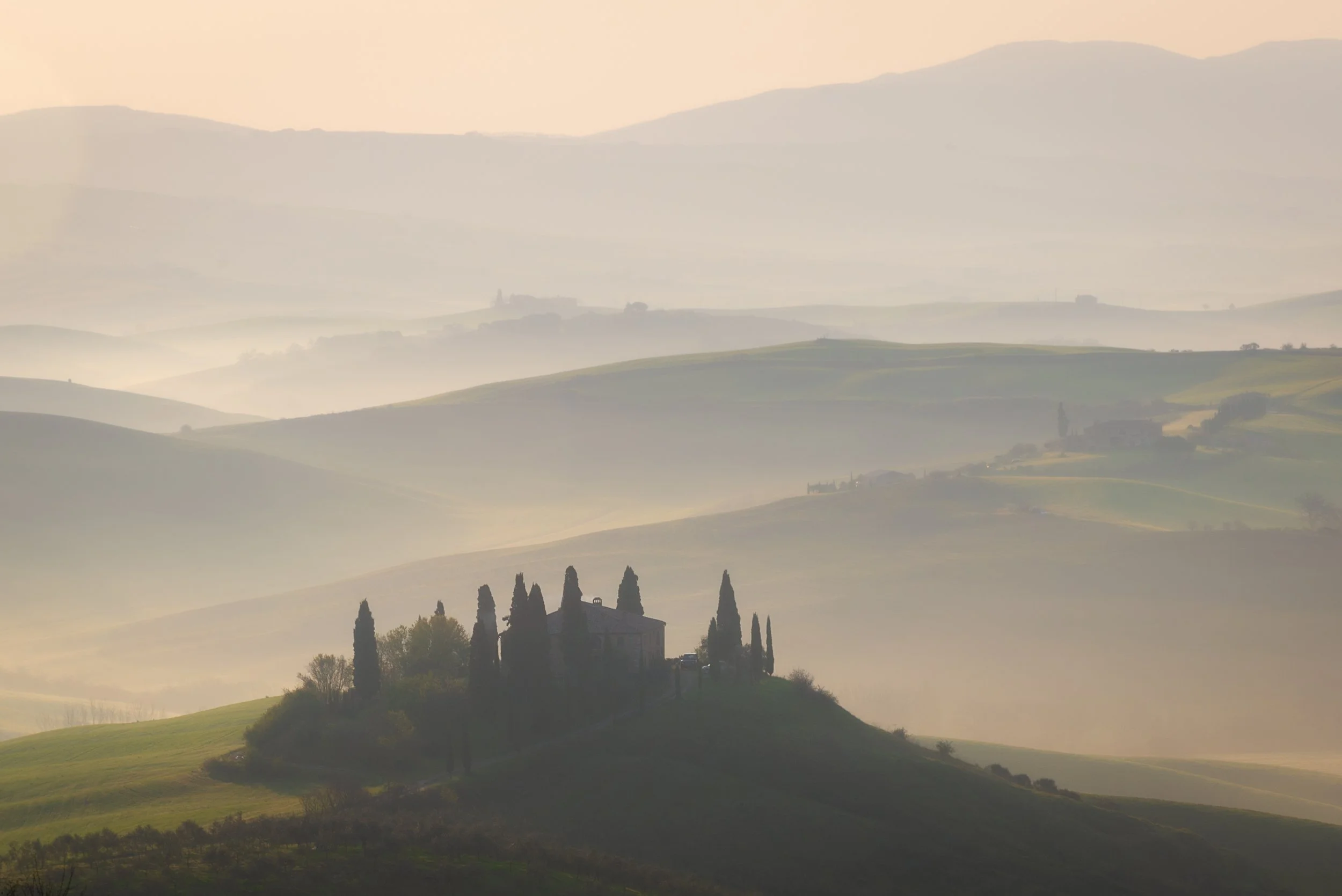 A misty landscape of rolling hills with a house surrounded by tall cypress trees in the foreground, and multiple layers of hills fading into the distance.