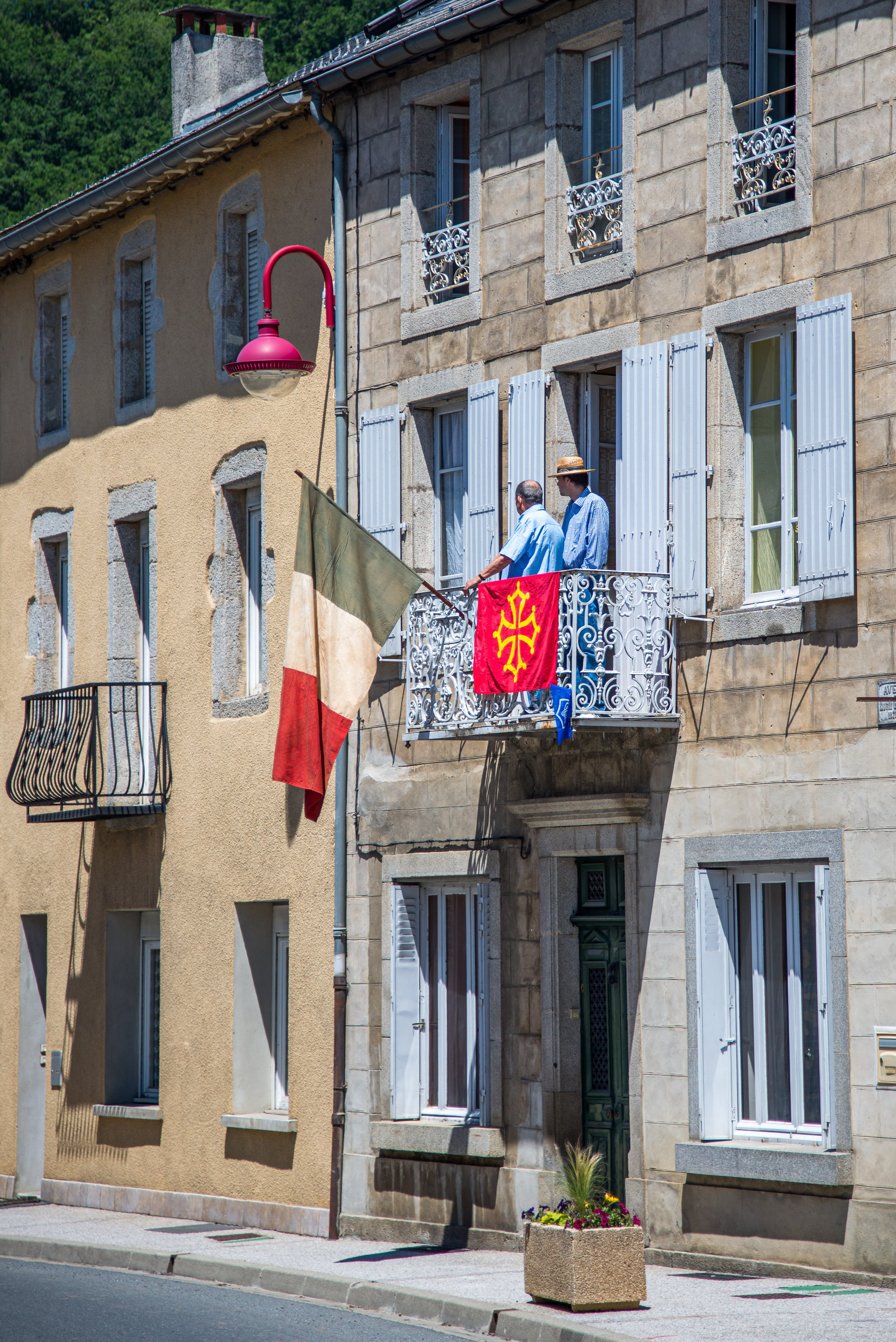 Two men on a small balcony of an old stone building, holding an Italian flag and a red banner with yellow symbols, with open blue shutters and a pink streetlamp.