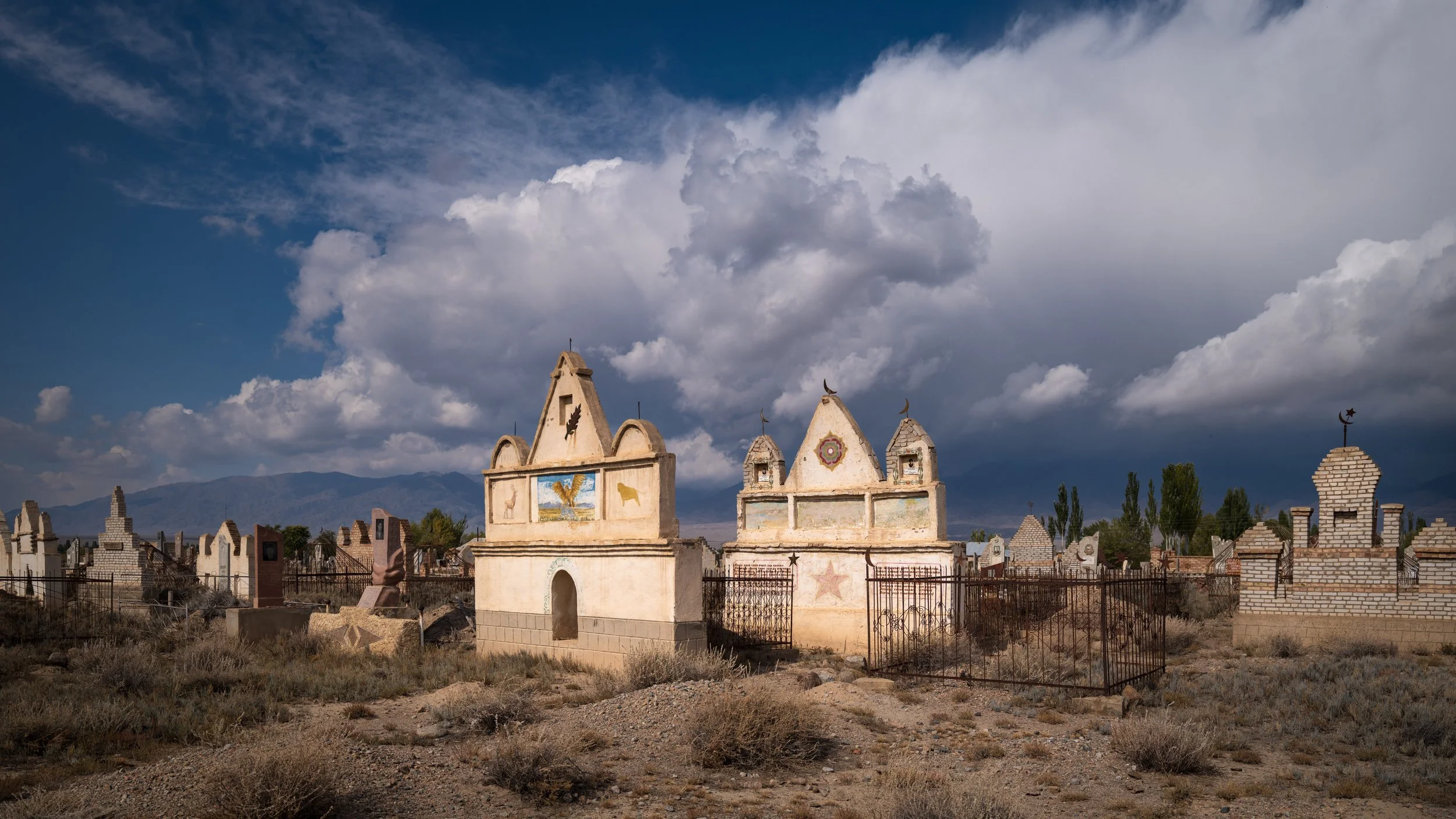 A desert cemetery with old, weathered tombstones and mausoleums under a cloudy sky, with mountains in the background.