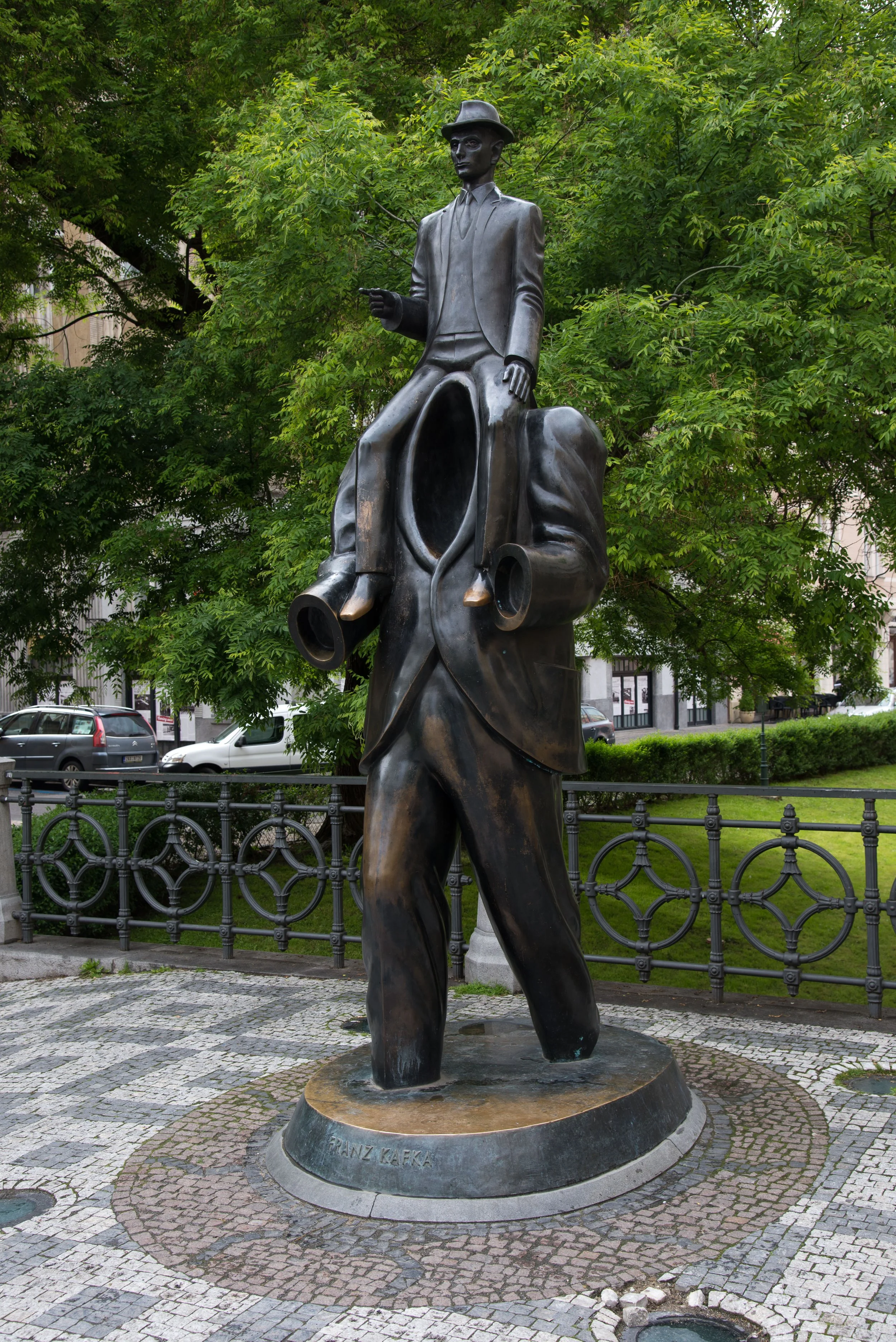 Bronze statue of a man in a suit and hat, sitting on a large speaker, holding a small object, with trees and parked cars in the background.