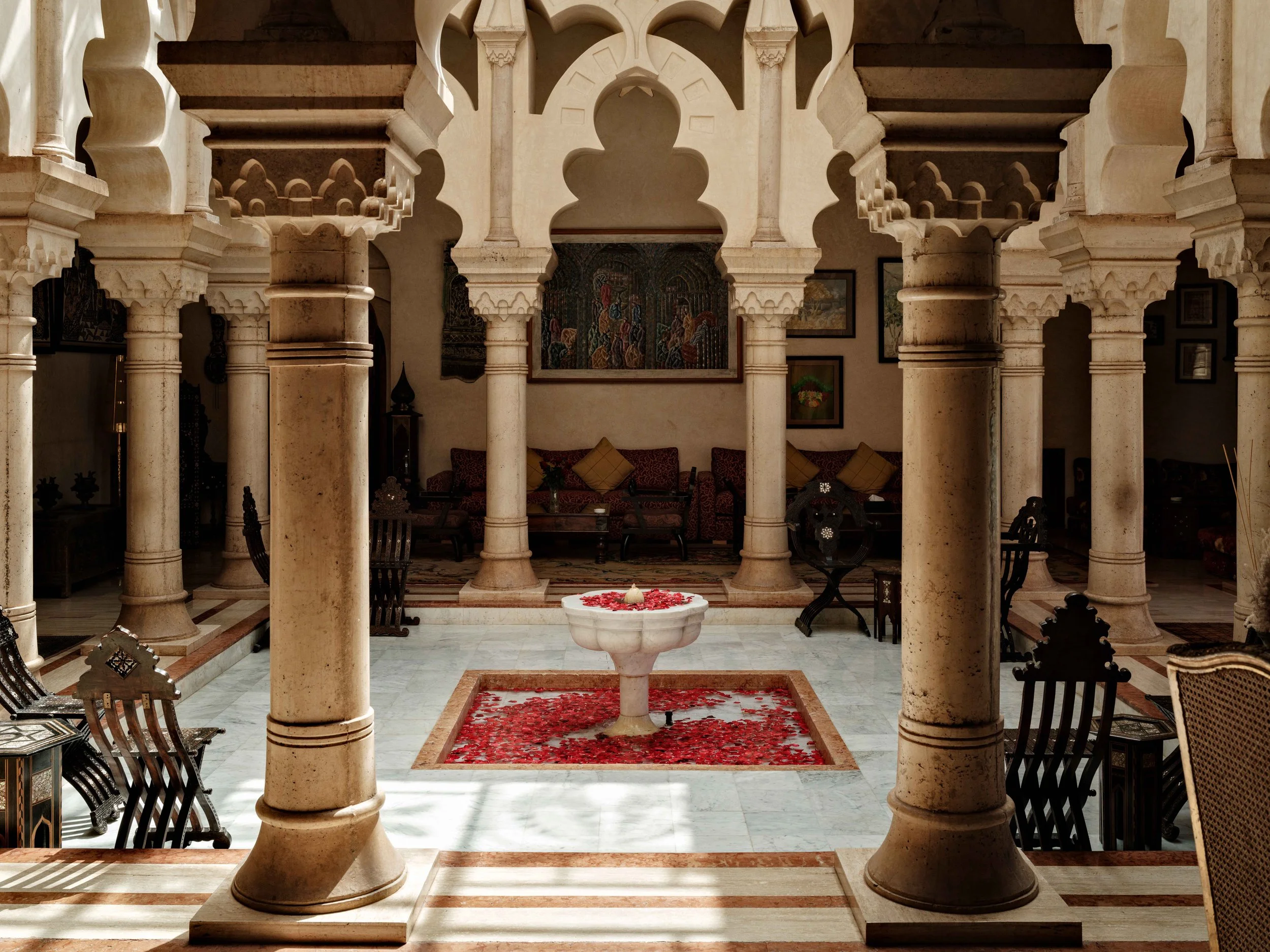 Interior view of a traditional Moroccan riad with ornate arches, stone columns, a central fountain with rose petals, surrounded by wooden chairs and sofas, and decorated with paintings on the walls.
