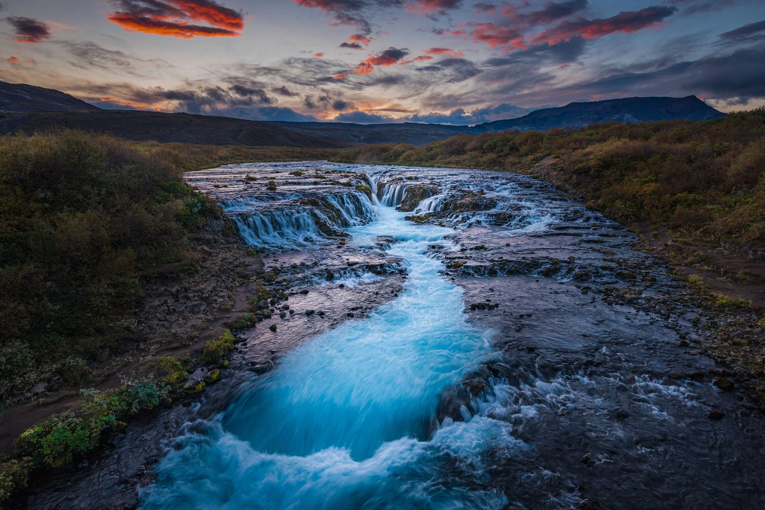 A river flowing through a rugged landscape at sunset, with a layered sky filled with orange, pink, and purple clouds and mountains in the distance.