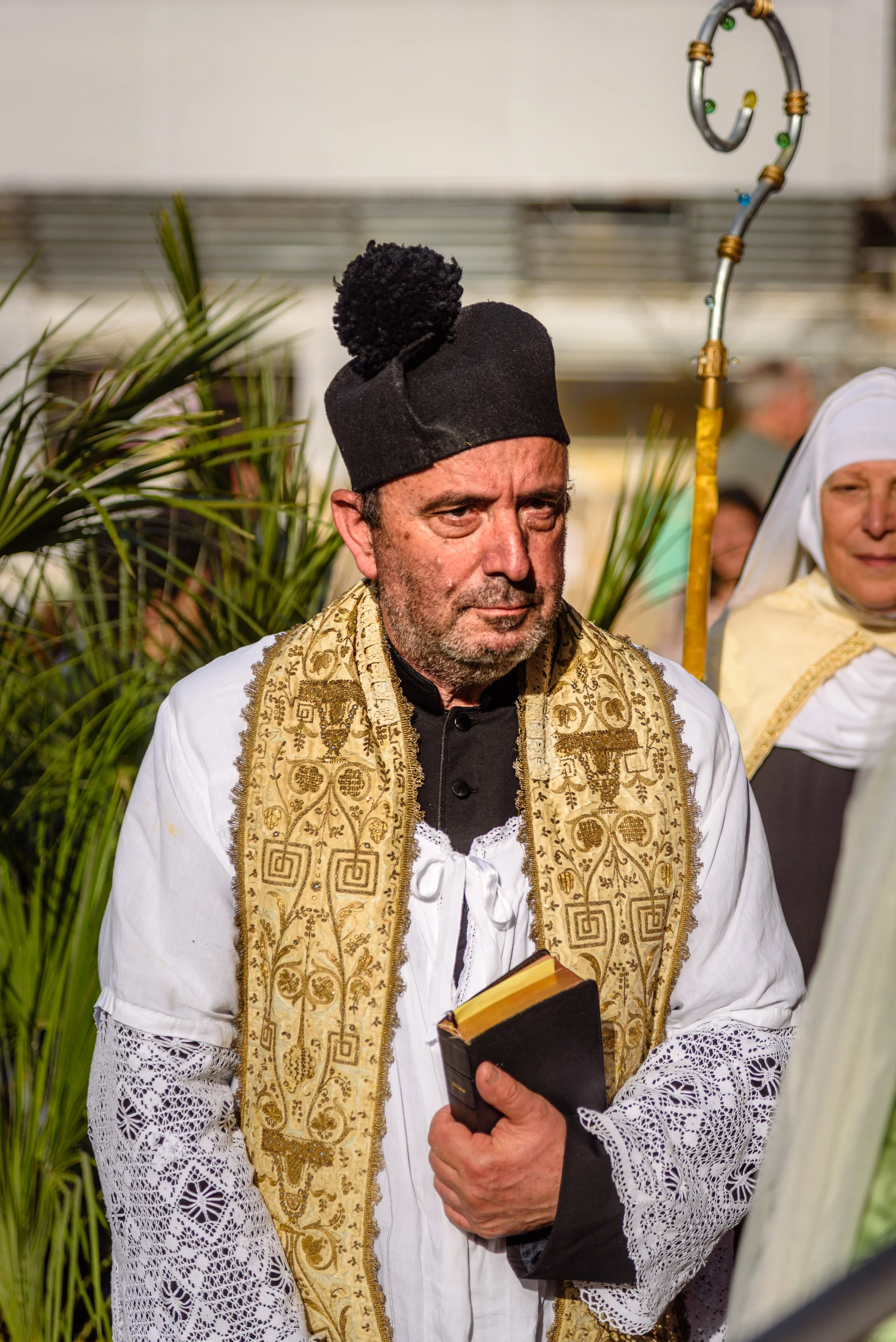 Man dressed in religious or ceremonial attire holding a book, wearing a black hat with a pom-pom, and a gold embroidered stole over a white garment, standing among other people in similar attire.