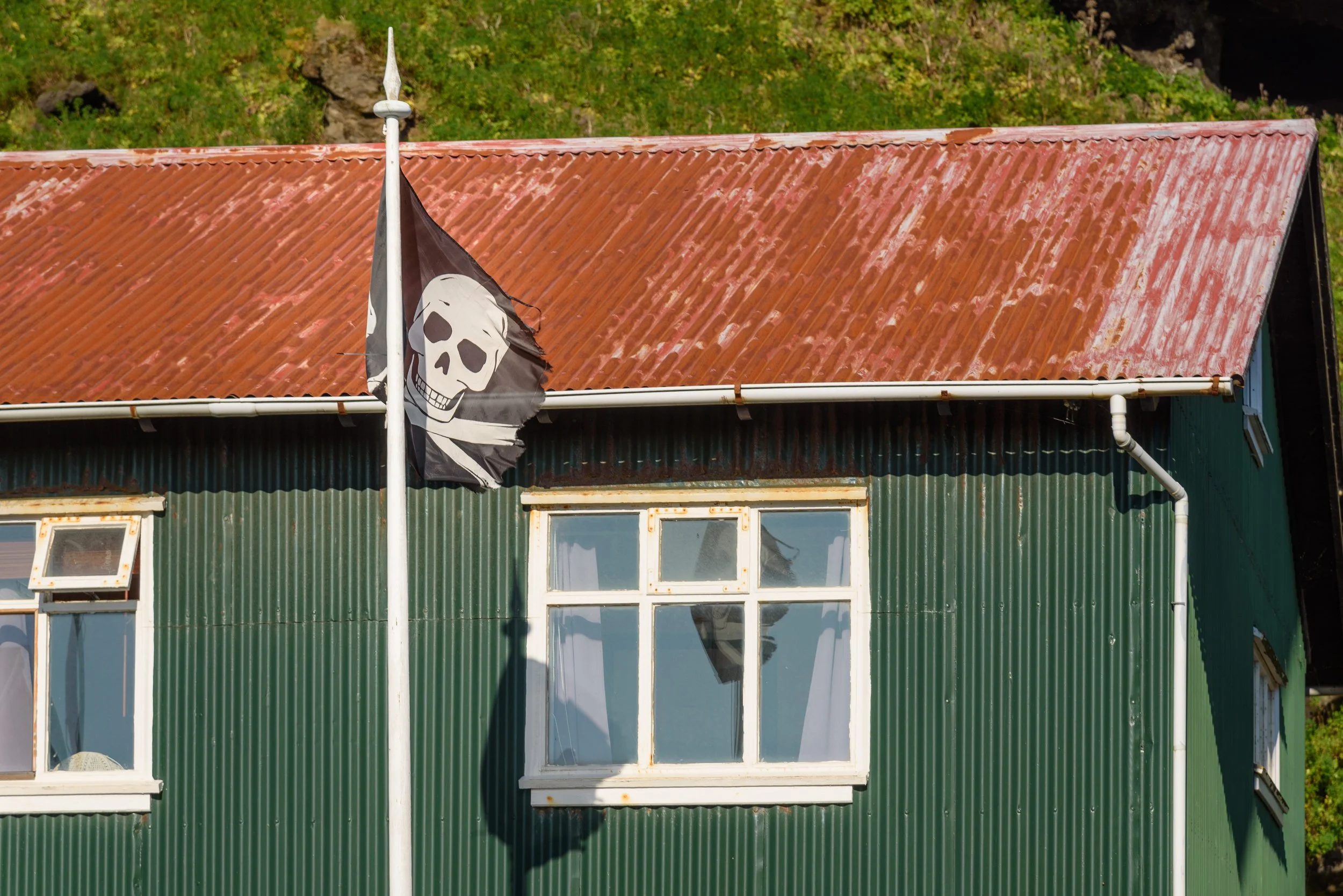 A green corrugated metal house with white-framed windows and a rusted red metal roof. A black pirate flag with a white skull and crossbones is flying on a flagpole in front of the house.