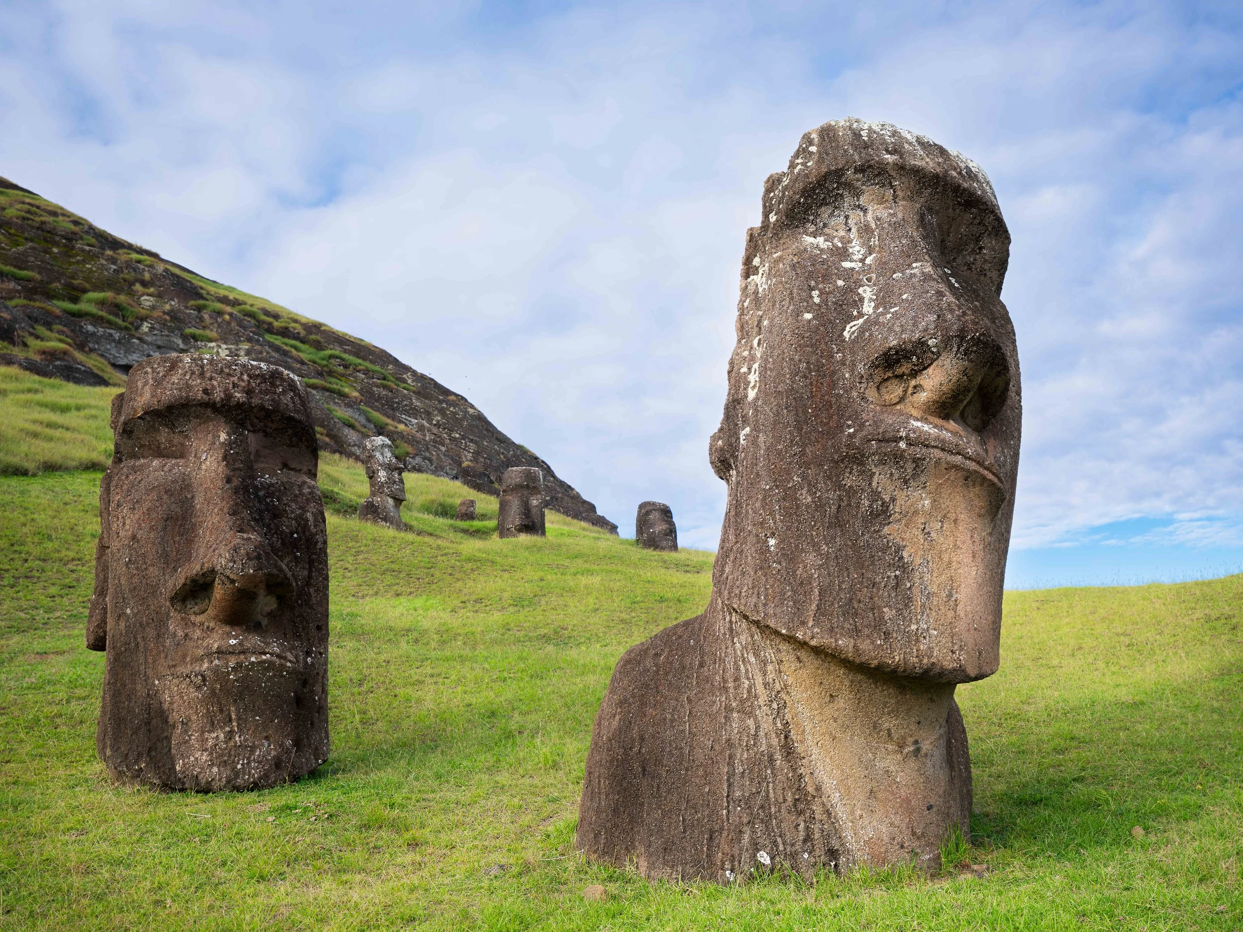 Multiple large stone Moai statues on a grassy hillside with a partly cloudy sky.