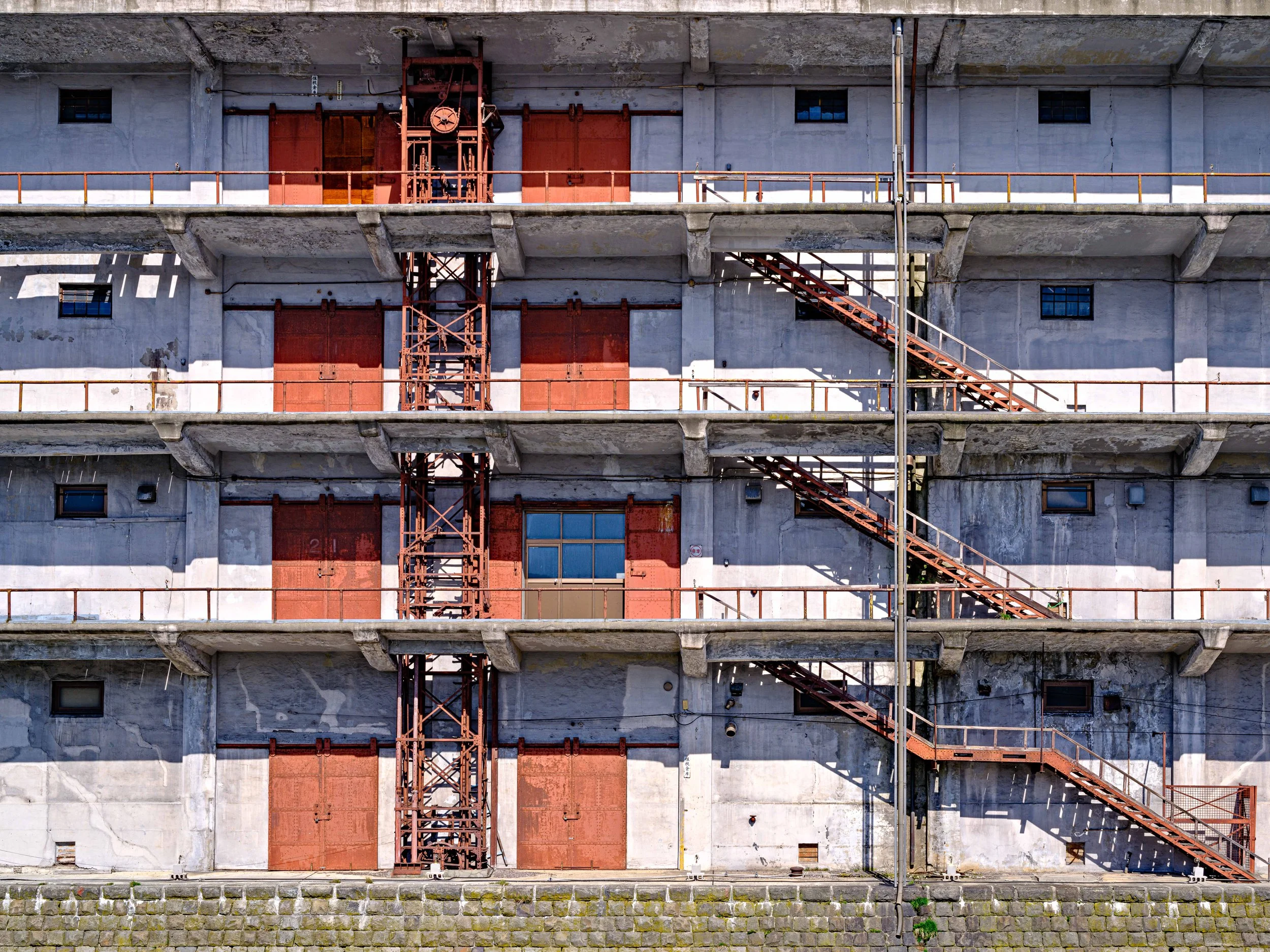 An exterior view of a concrete building with red metal doors and stairs, and a central vertical metal staircase.