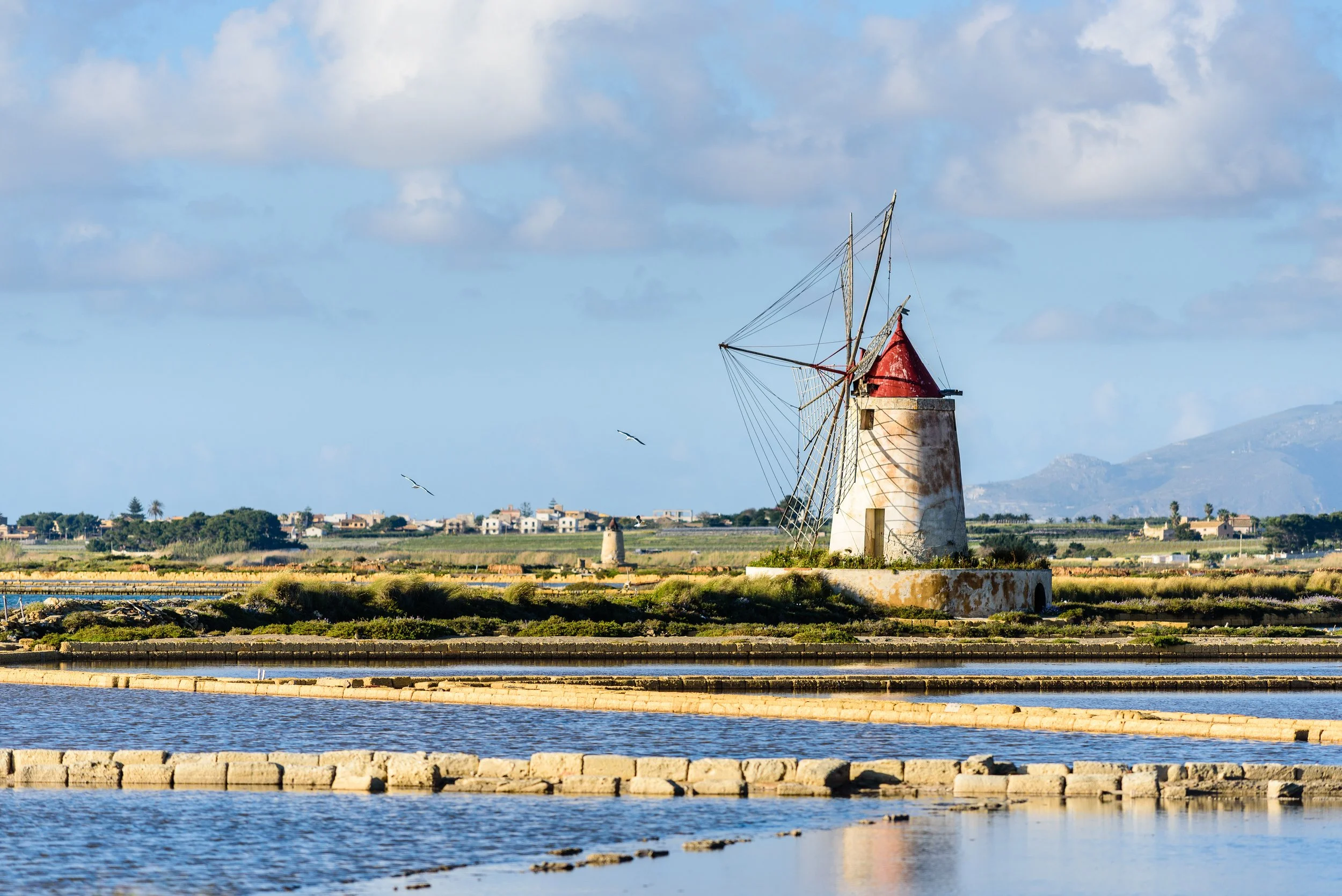 A scenic view of a small windmill with a red conical roof, situated near salt ponds, with a few seagulls flying in a partly cloudy sky, and a distant village and mountains in the background.