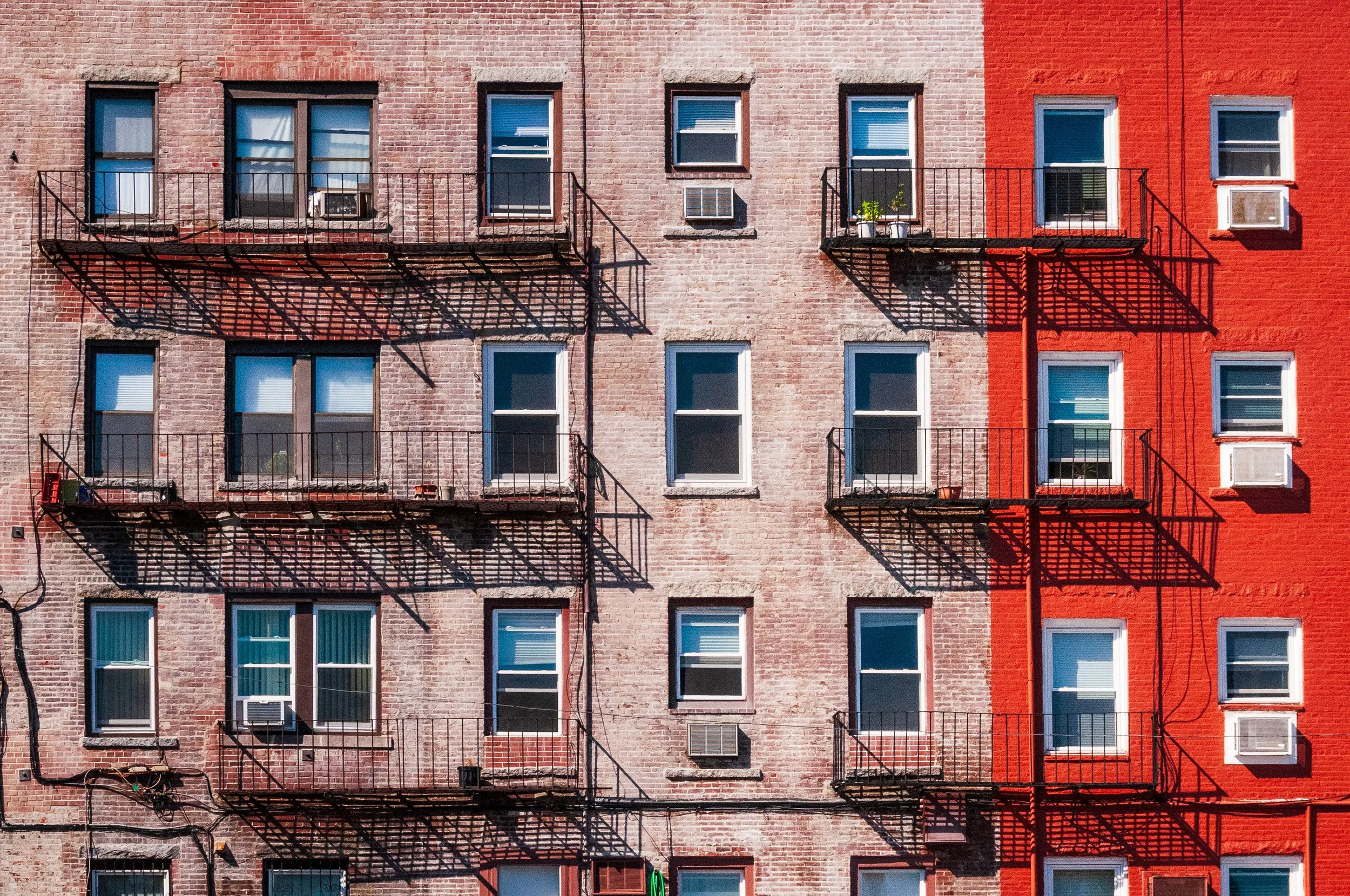 The exterior of a brick apartment building with fire escapes on each floor, casting shadows on the facade. The building has a section painted red on the right. Several windows with air conditioning units and a potted plant are visible.