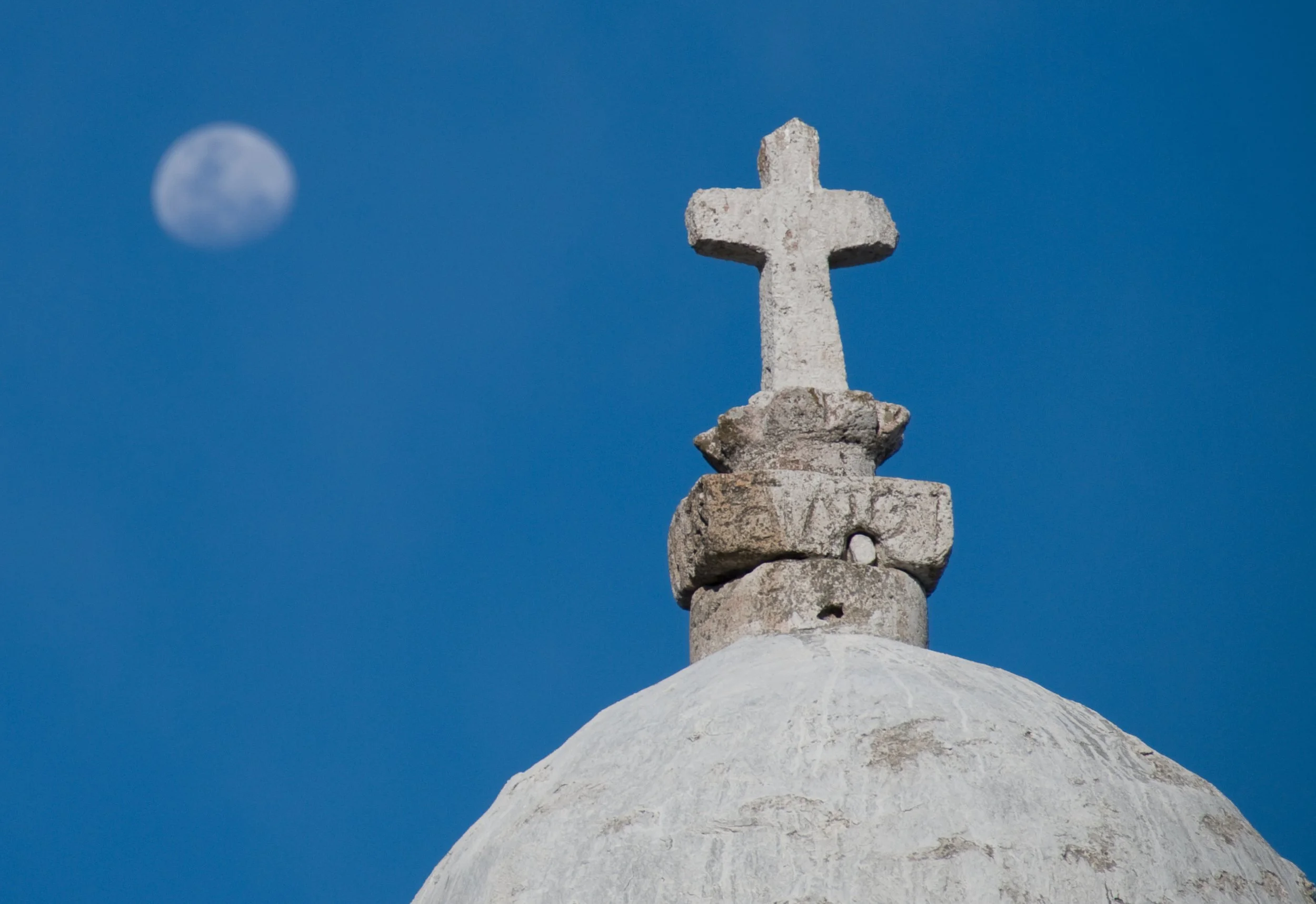 A stone cross on top of a white dome building against a blue sky with visible moon in the background