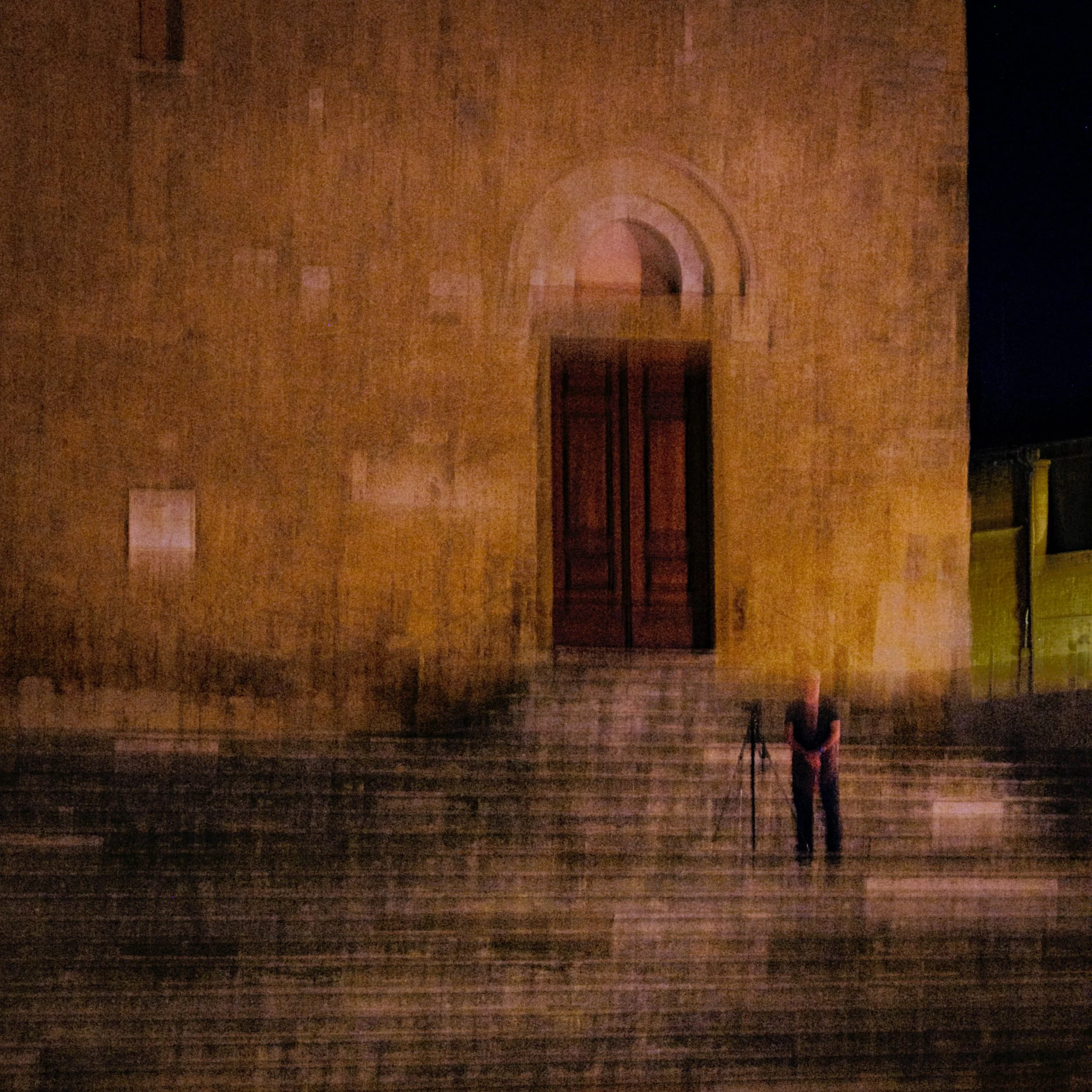 A person with a camera on a tripod standing on steps in front of a large, illuminated building at night.