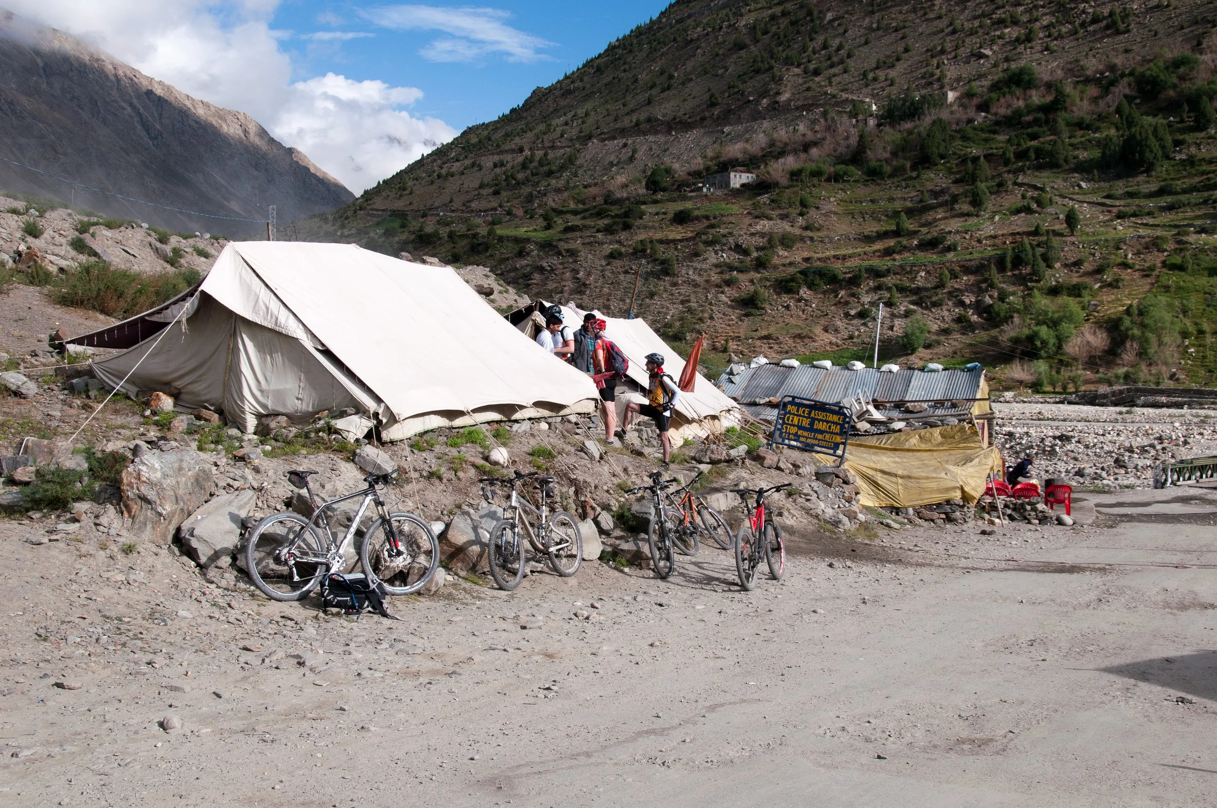 Group of cyclists resting and socializing near tents on a rocky mountain terrain with hills and sparse trees in the background.