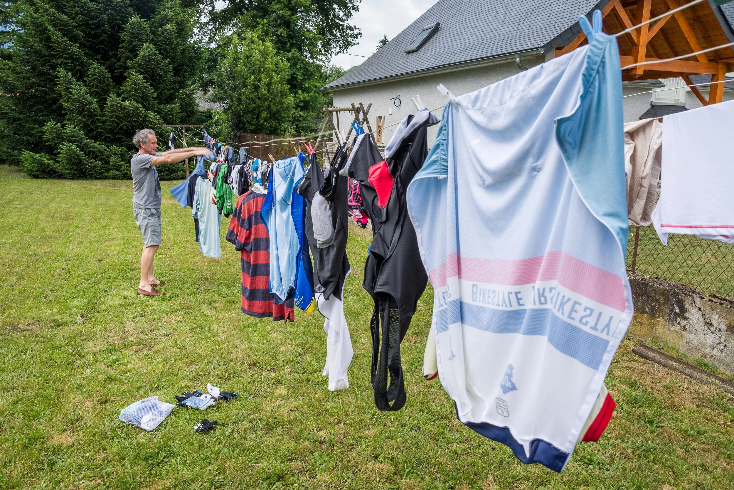 An elderly man hanging clothes on a clothesline outdoors in a backyard with green grass, trees, and a house.
