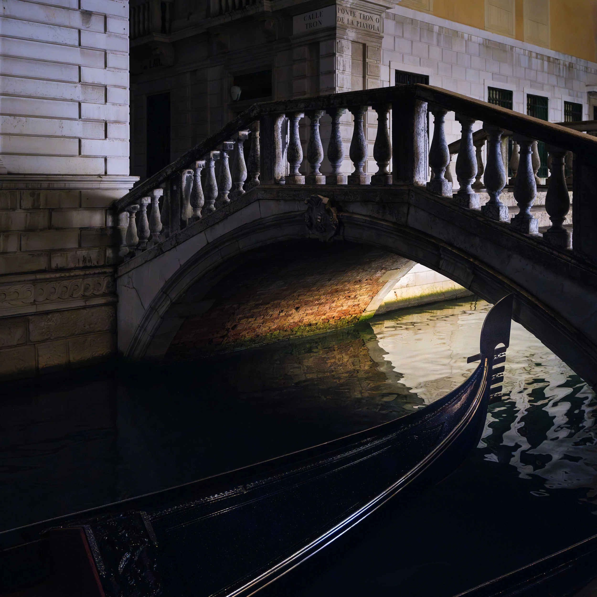 Nighttime view of a small stone bridge over a canal in Venice, Italy, with a gondola moored beneath it. The surrounding buildings are illuminated softly.