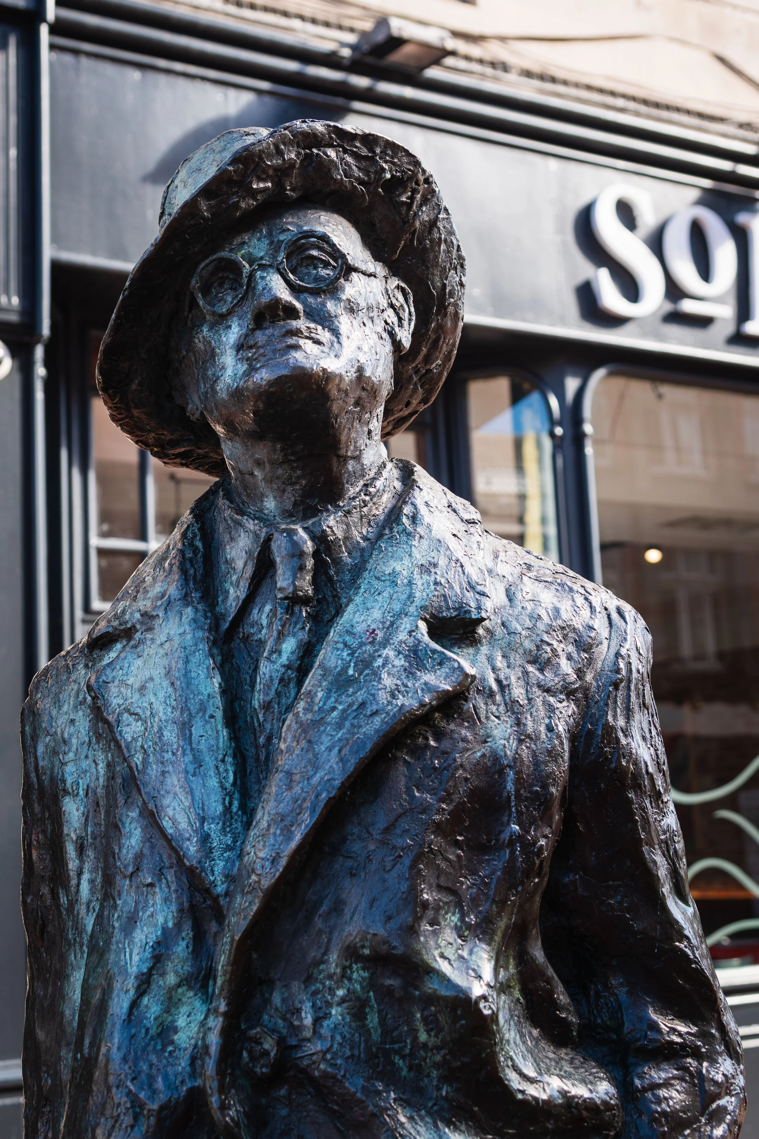 A bronze statue of a man wearing glasses and a hat, dressed in a suit and tie, standing in front of a building with signs and windows.