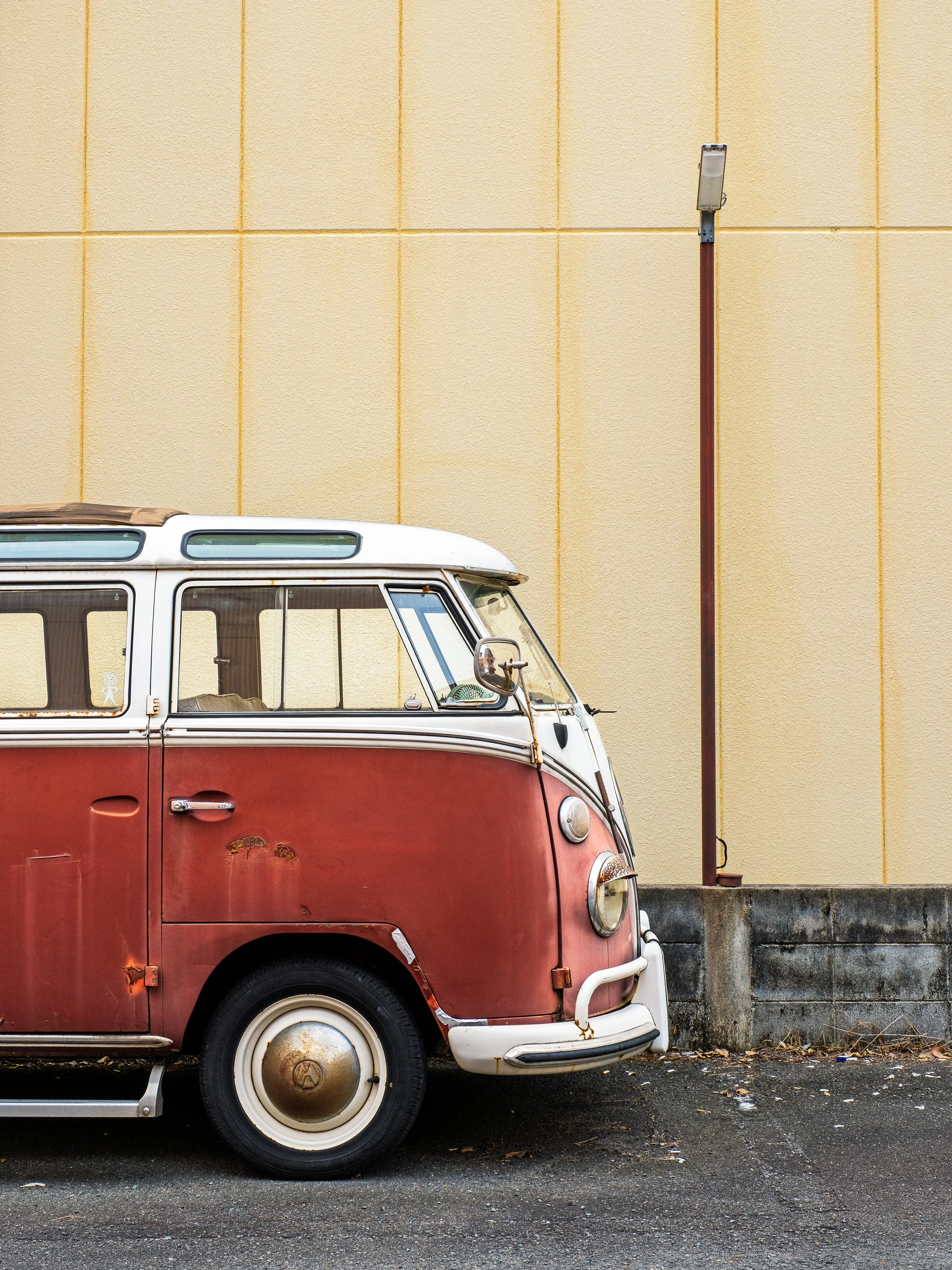 A vintage red and white Volkswagen camper van parked on a street.next to a yellow wall and a tall streetlamp.