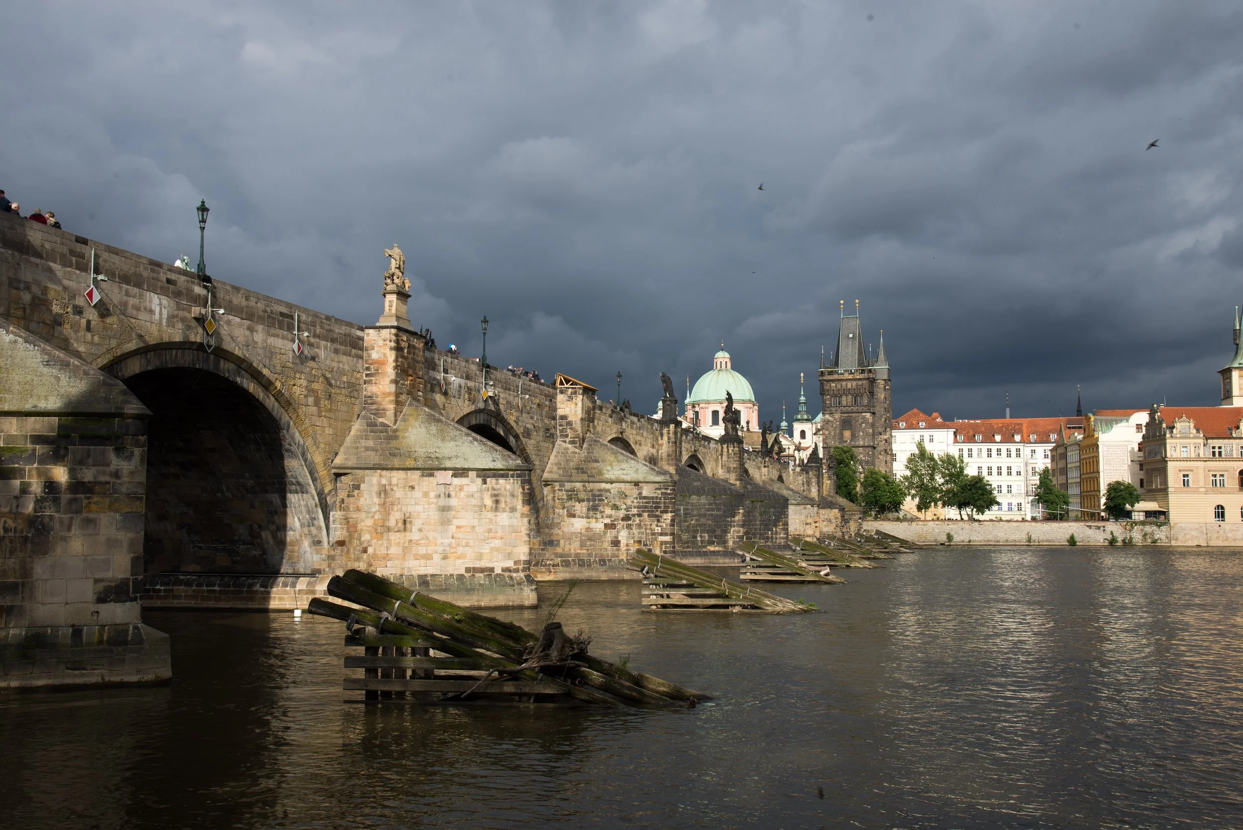 View of Charles Bridge in Prague, Czech Republic, with dark stormy clouds overhead and historic buildings along the riverbank.