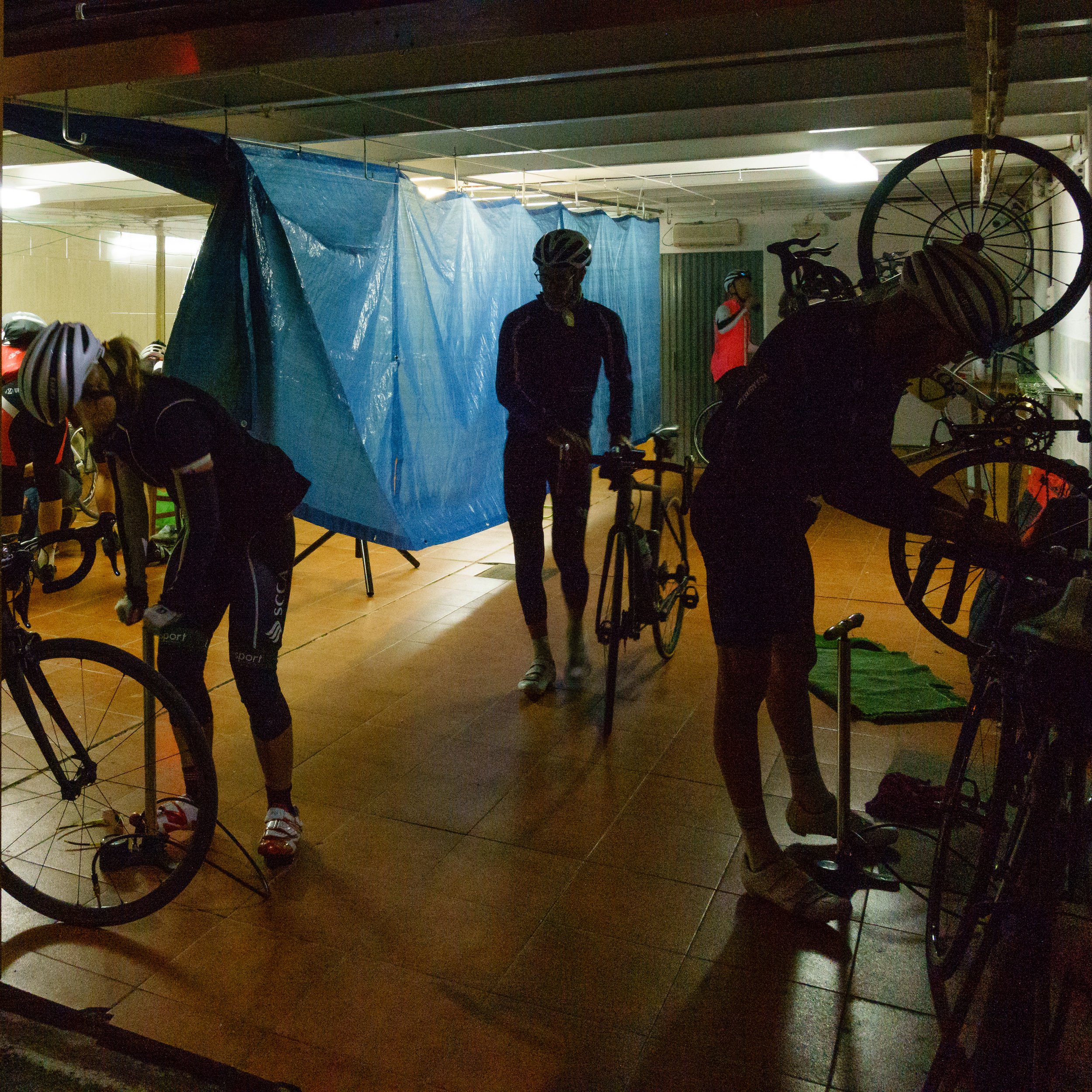Cyclists preparing in a dimly lit indoor space, with some adjusting bicycles and others standing near a blue tarp and various cycling gear.