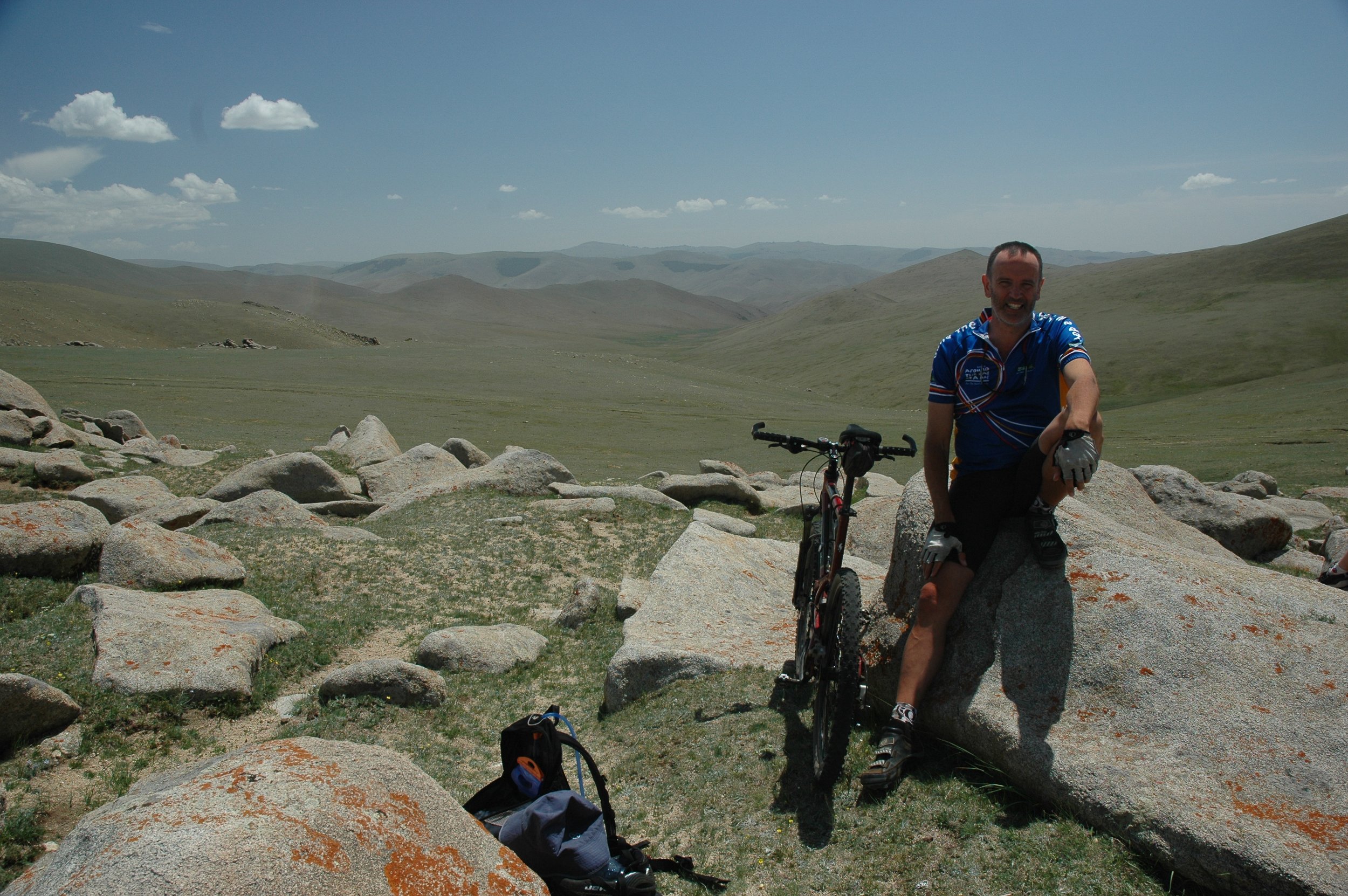 A man in cycling gear smiling while sitting on a large rock next to a mountain bike, in a vast, open, grassy landscape with rolling hills under a partly cloudy sky.