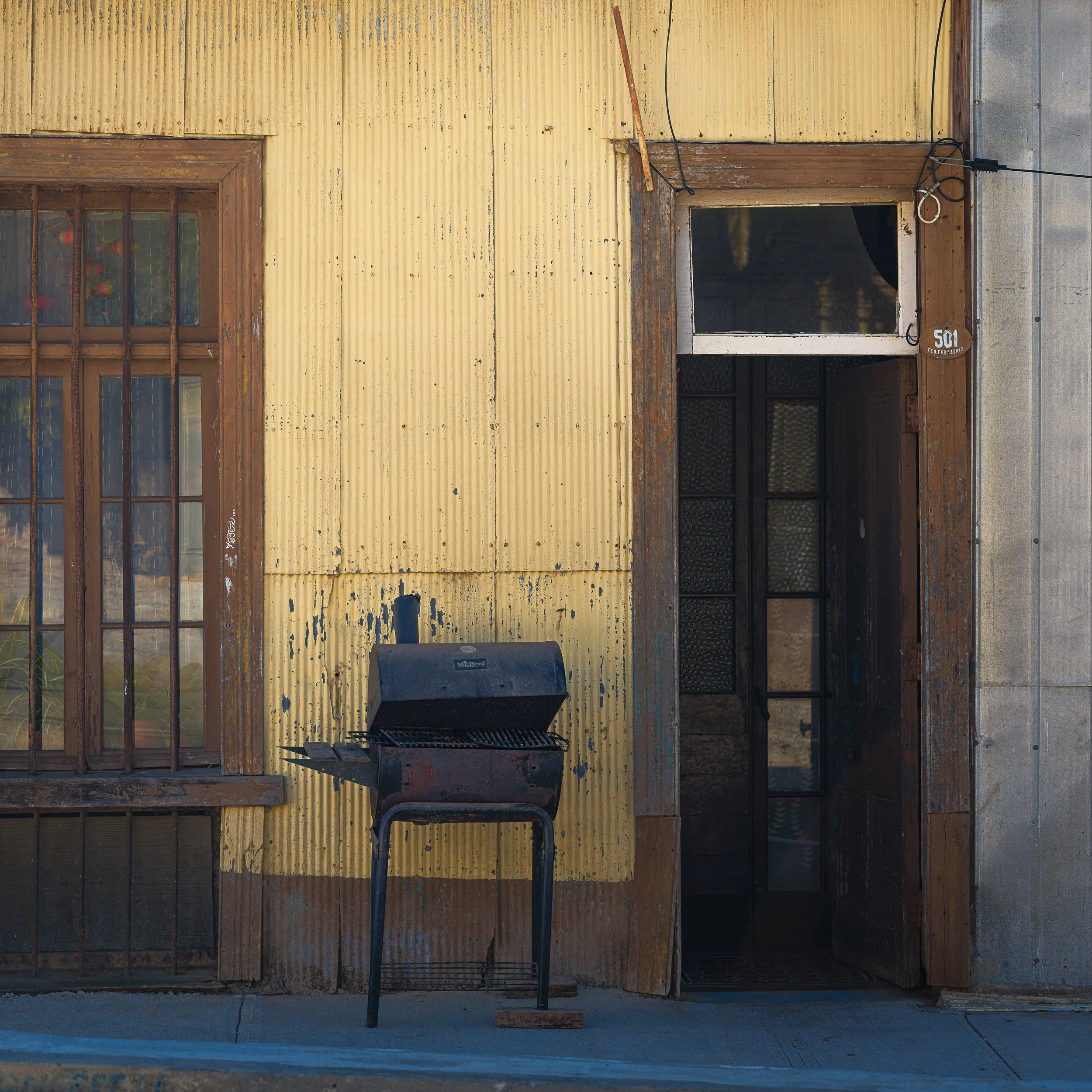 An exterior view of a rustic building with a yellow corrugated metal wall, a window with wooden frames and iron bars, a partially open door, and a black barbecue grill placed on the sidewalk.