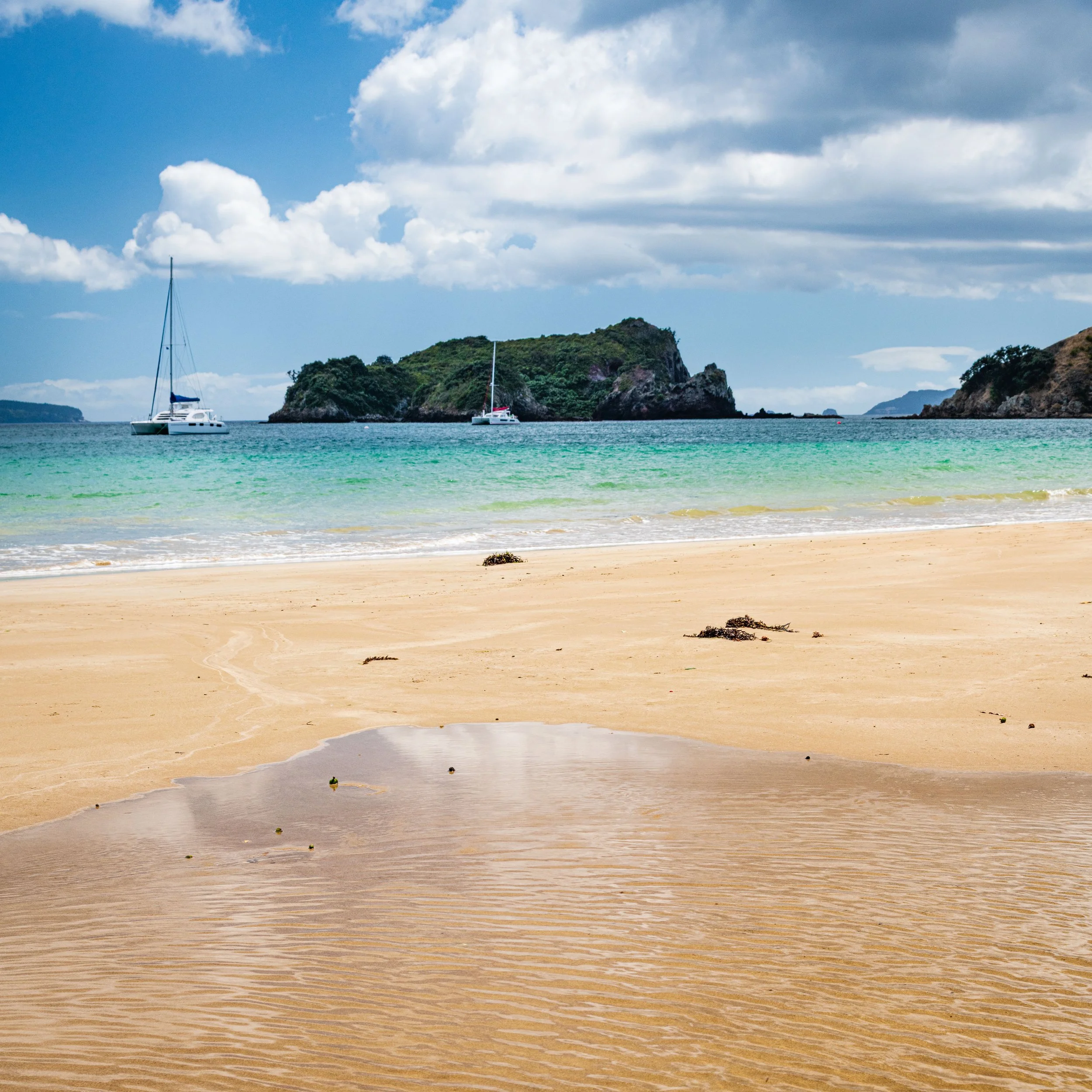 Sandy beach with small pools of water, sailboats anchored in calm water, green islands in the background, cloudy sky.