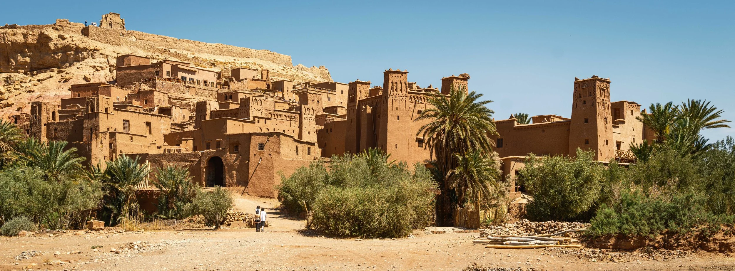 Traditional mud-brick village with multi-story buildings on a hillside, palm trees and desert landscape in the foreground under a clear blue sky.