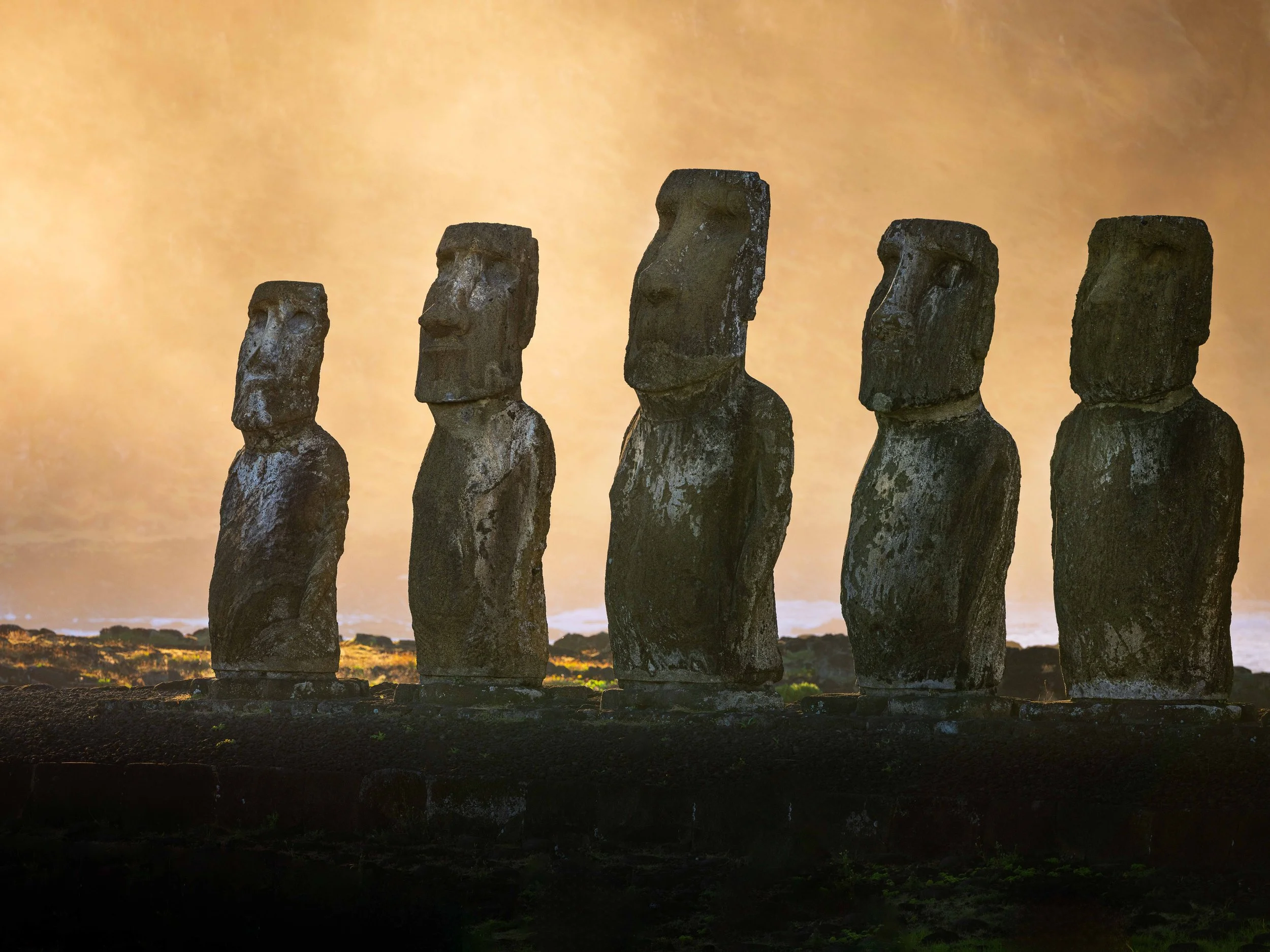 Five Moai statues standing on a stone platform at sunset with a warm, glowing sky in the background.