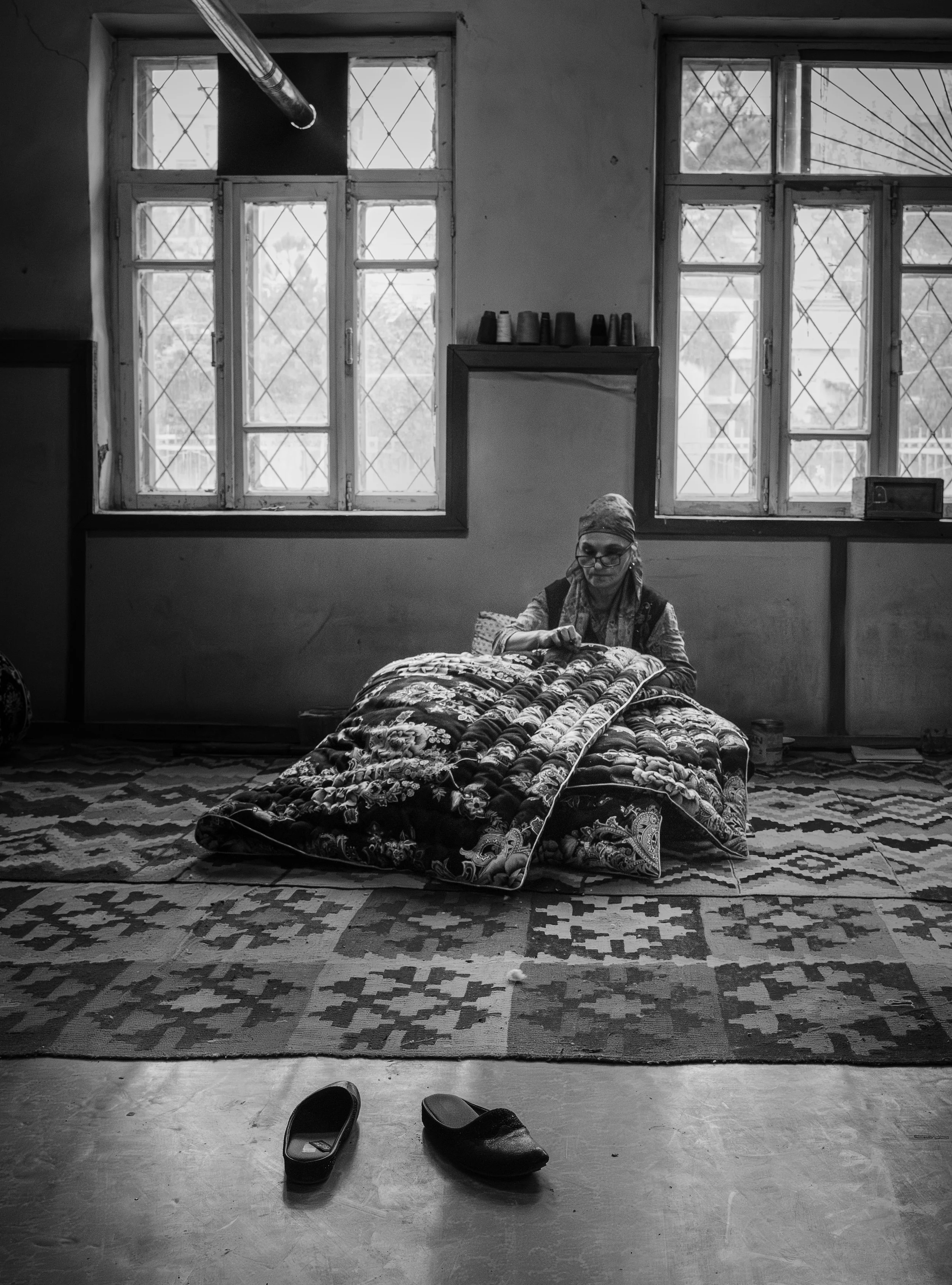 A woman sitting on a the floor, sewing a large quilted blanket inside a room with large windows, with slippers on the floor in the foreground.
