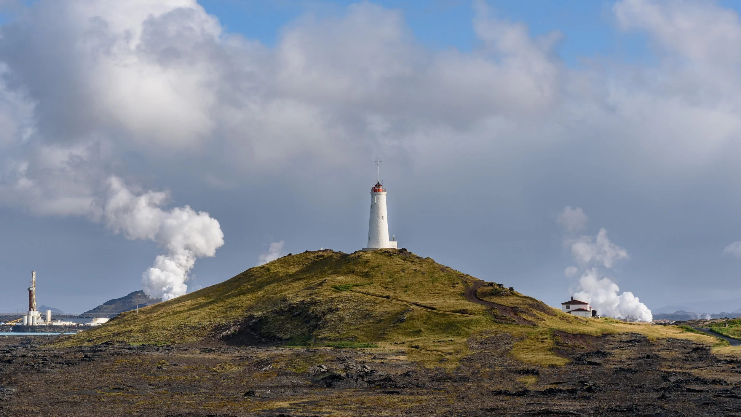 Lighthouse on a grassy hill with smoke billowing from industrial chimneys in the background and a partly cloudy sky overhead.