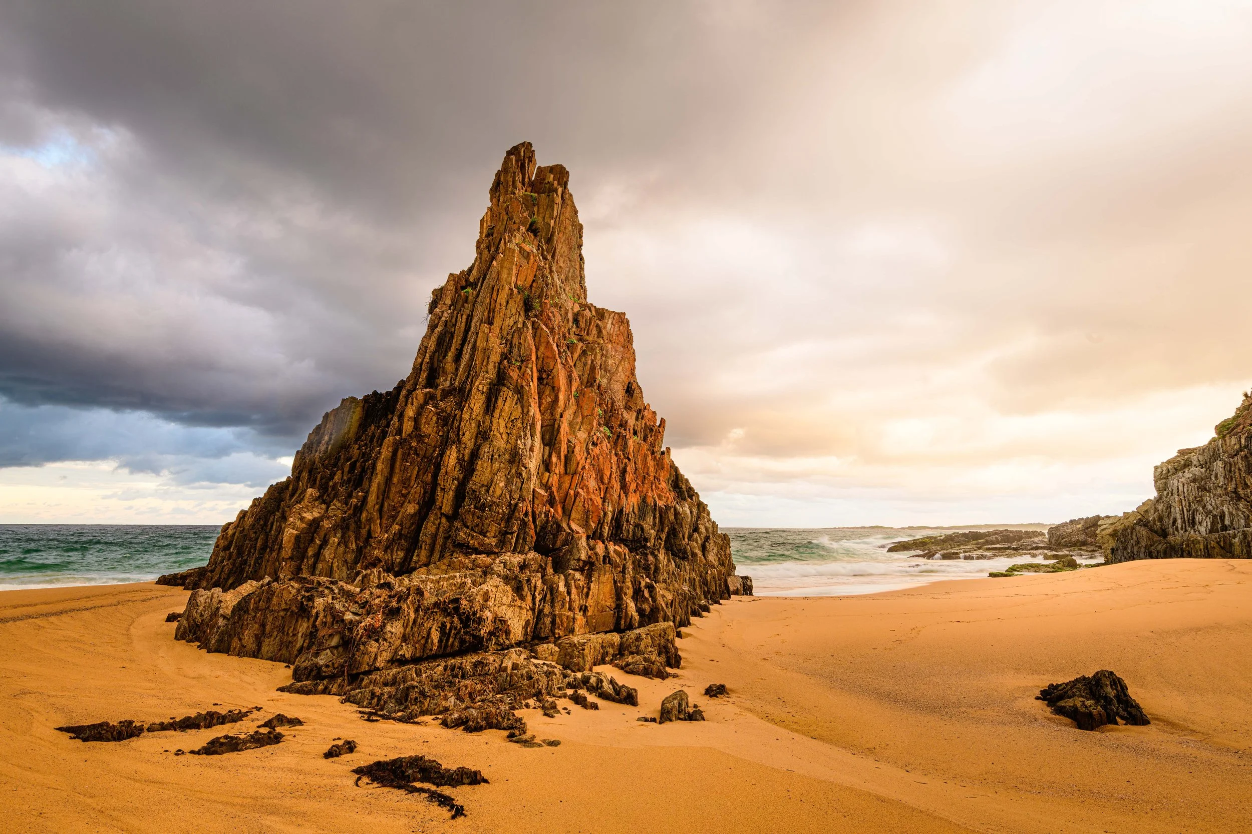 Large jutting rock formation erupts from the textured sand on Gillard's Beach in late afternoon sunshine Mimosa Rocks National Park Tathra NSW | Brisbane landscape and travel photographer Ralph McConaghy