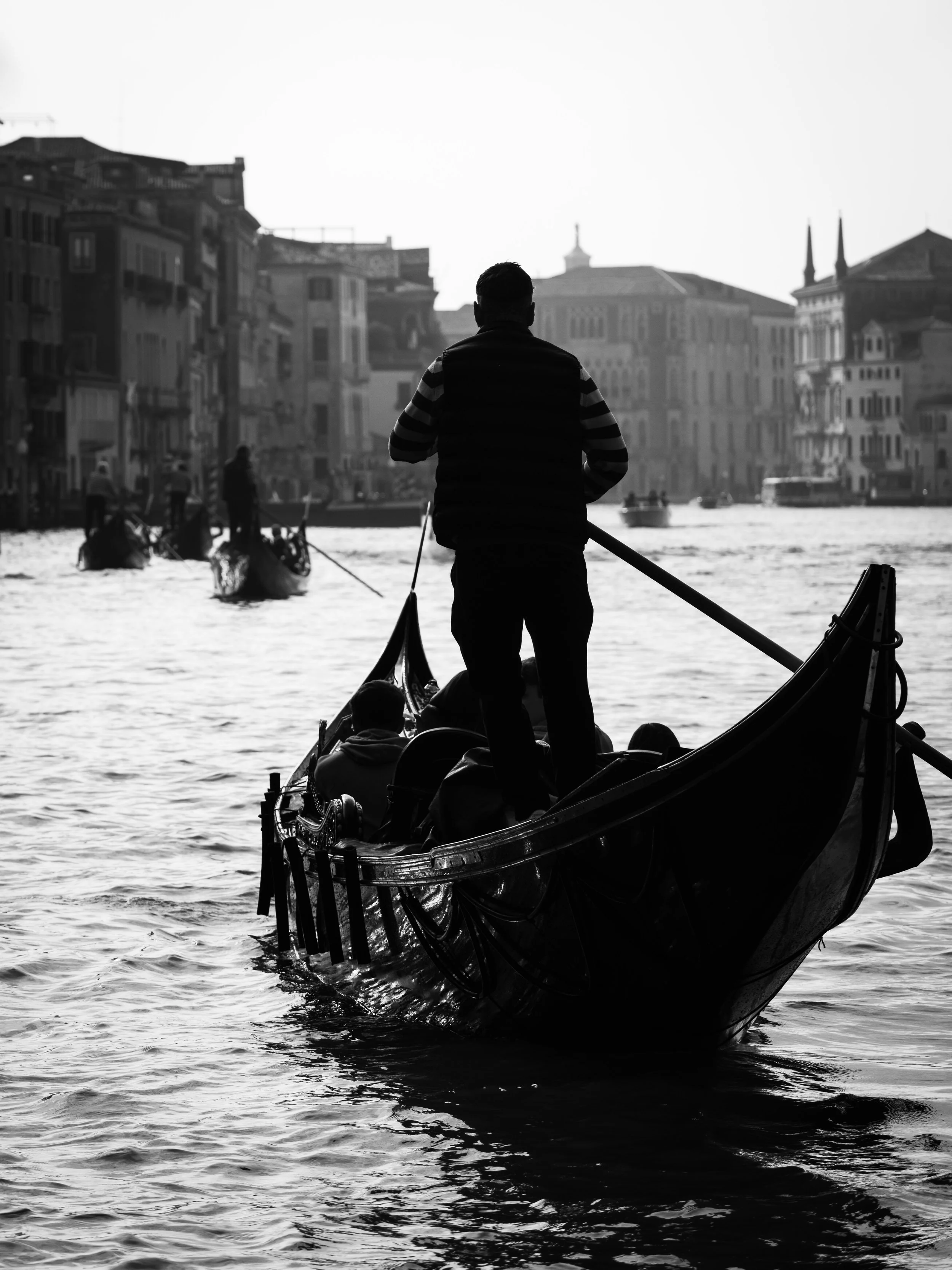 A black and white photo of a man standing on a gondola in a canal, with other gondolas and historic buildings in the background, typical of Venice, Italy.