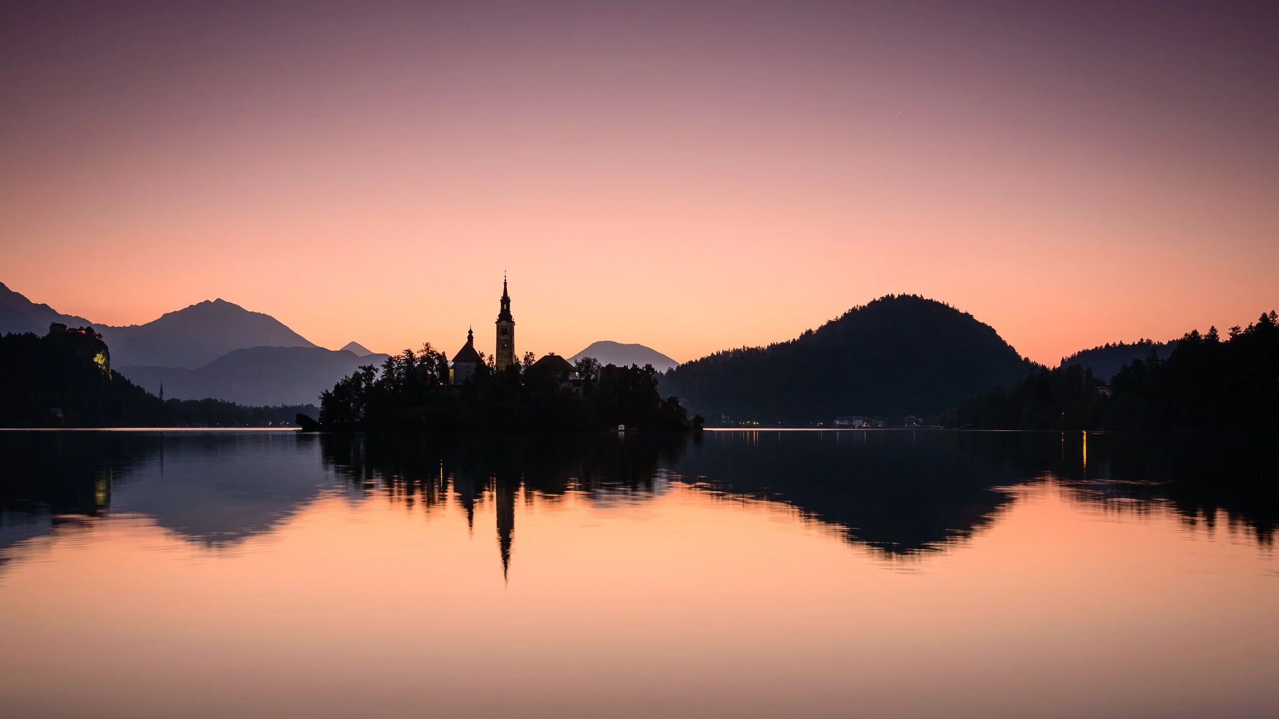 A tranquil lake at sunset with an island featuring a church, mountains in the background, and the sky transitioning from pink to purple, reflected in the water.
