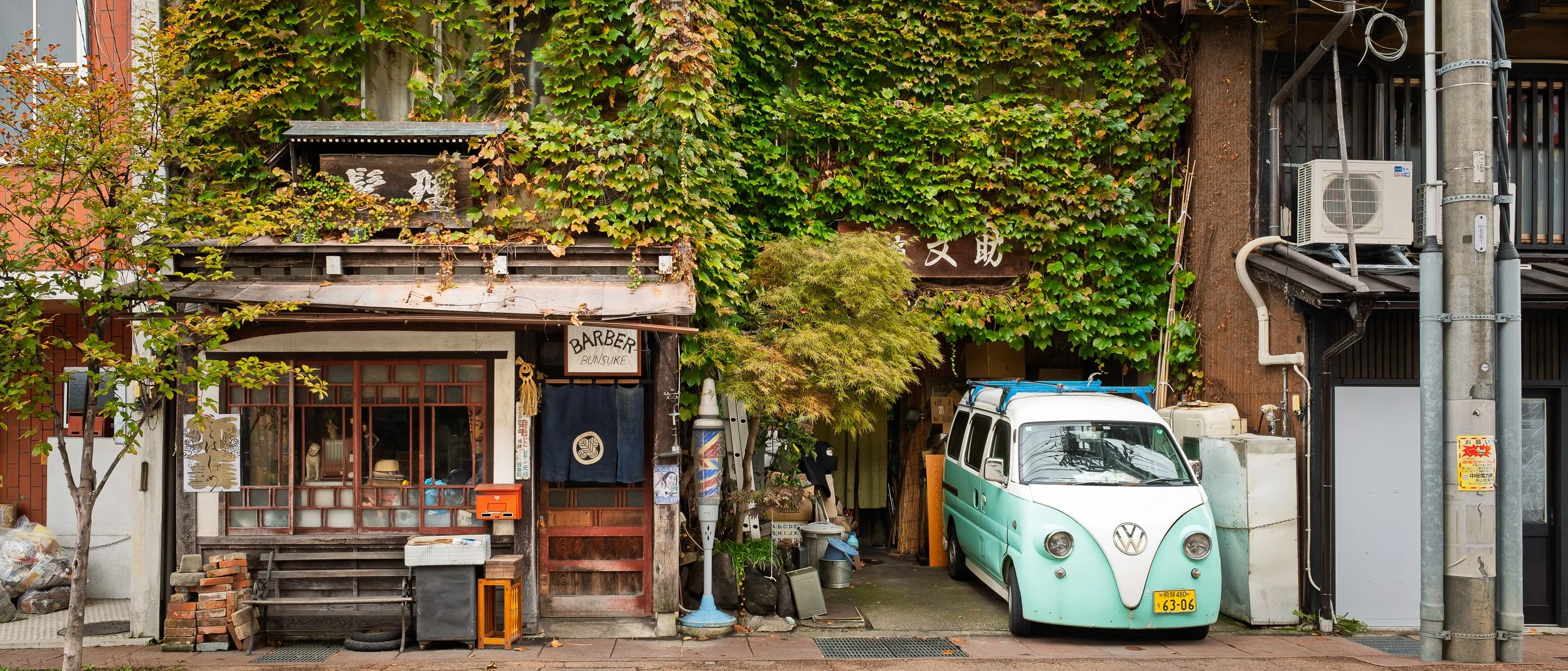 A Japanese storefront with a mix of traditional and modern elements, including a barber shop sign, lush green ivy covering the building, a small tree, and a retro Volkswagen van parked on the street. There are various items outside, such as a barber 