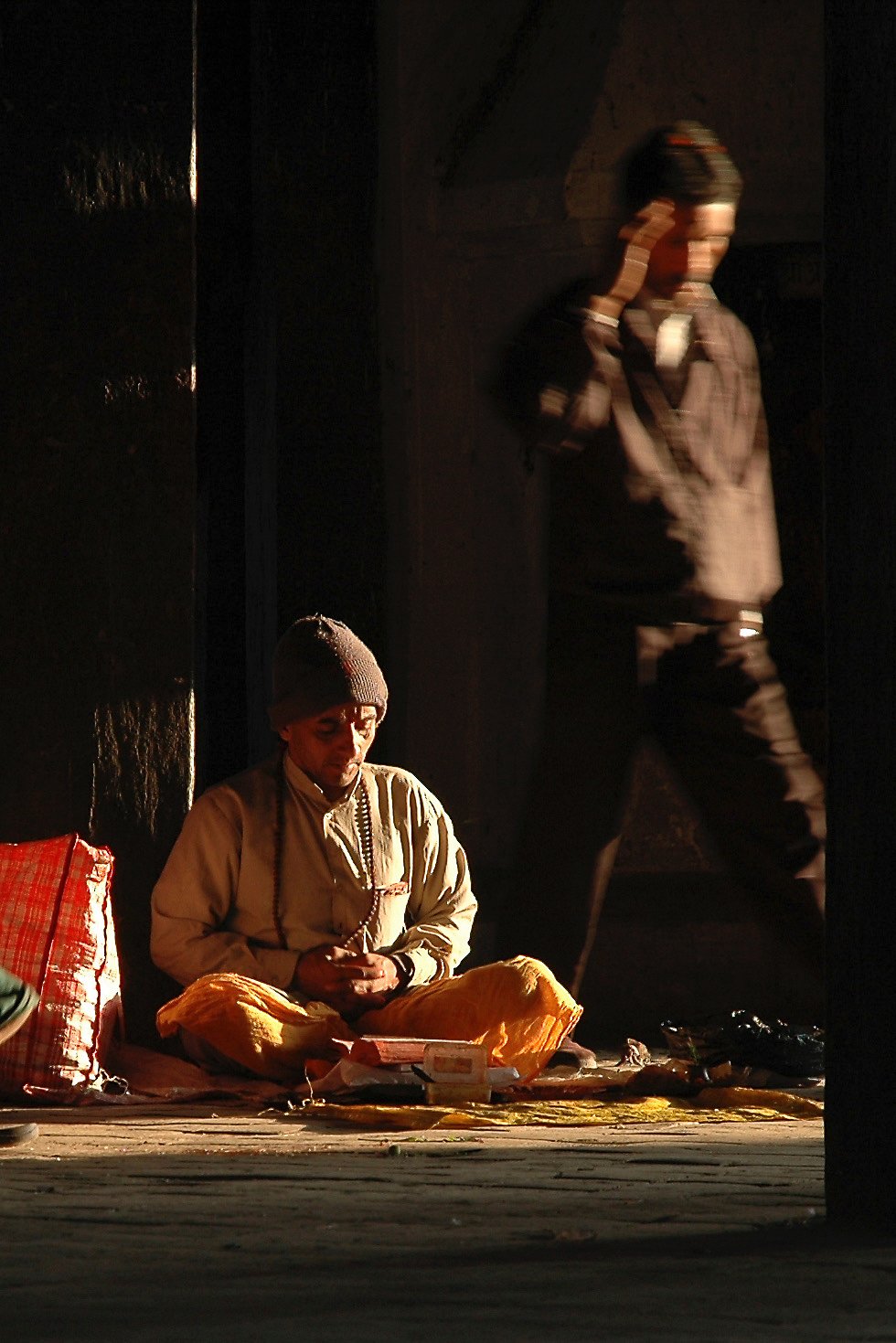 A person sitting cross-legged on the ground, wearing yellow pants, a beige jacket, and a gray beanie, appears to be praying or meditating inside a dimly lit space. Another person, dressed in black and gray, stands nearby with one hand on their head, 