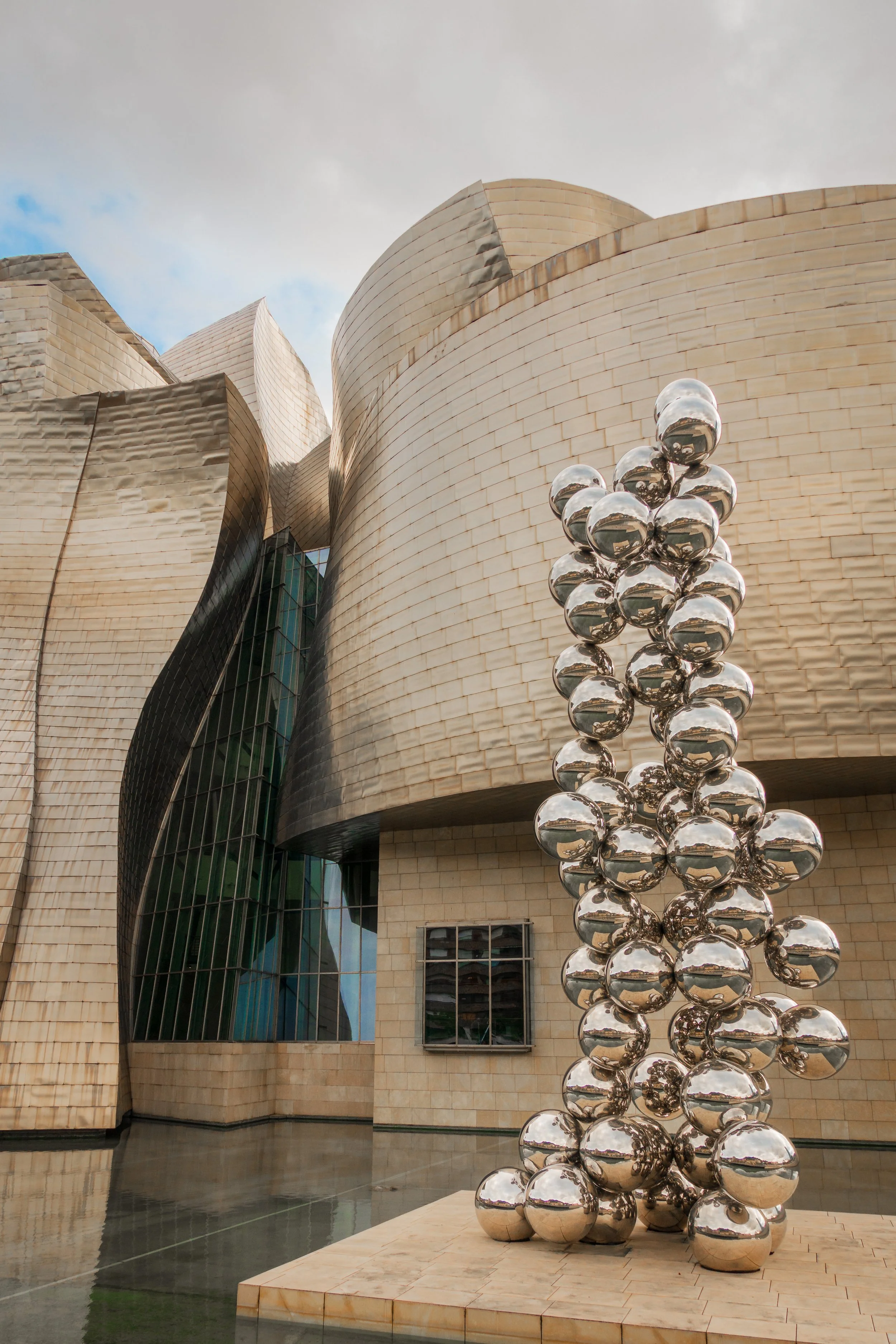 A modern building with irregular, metallic exterior panels and a glass section, alongside a sculpture made of reflective silver spheres stacked in a vertical formation.