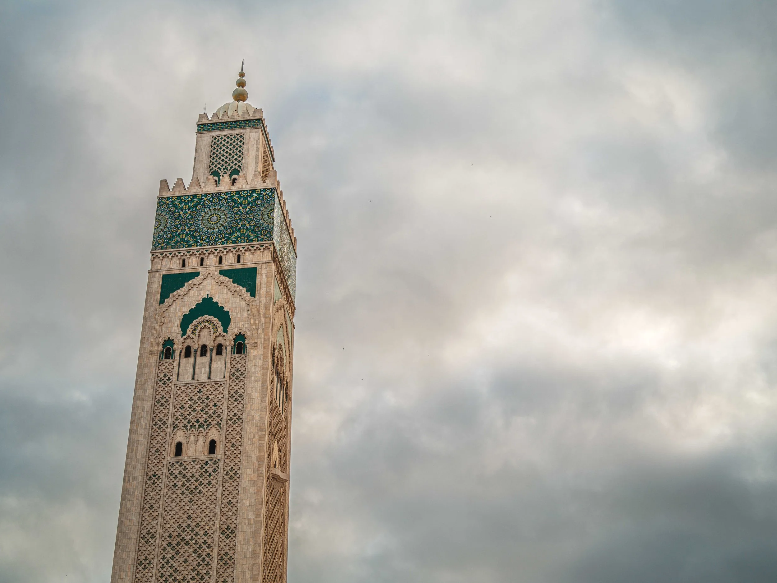 Near the top of a tall minaret with intricate tile work and decorative details, weathered clouds in the sky.