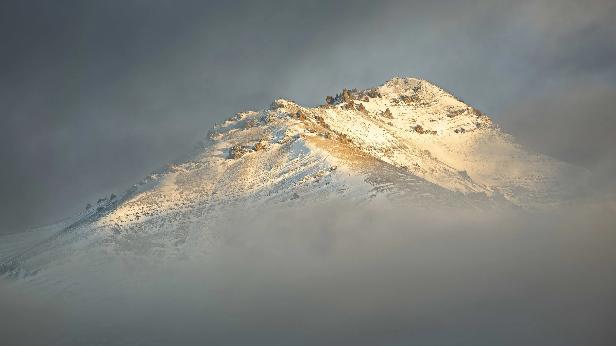 Snow-covered mountain peaked above clouds with dark sky in background.