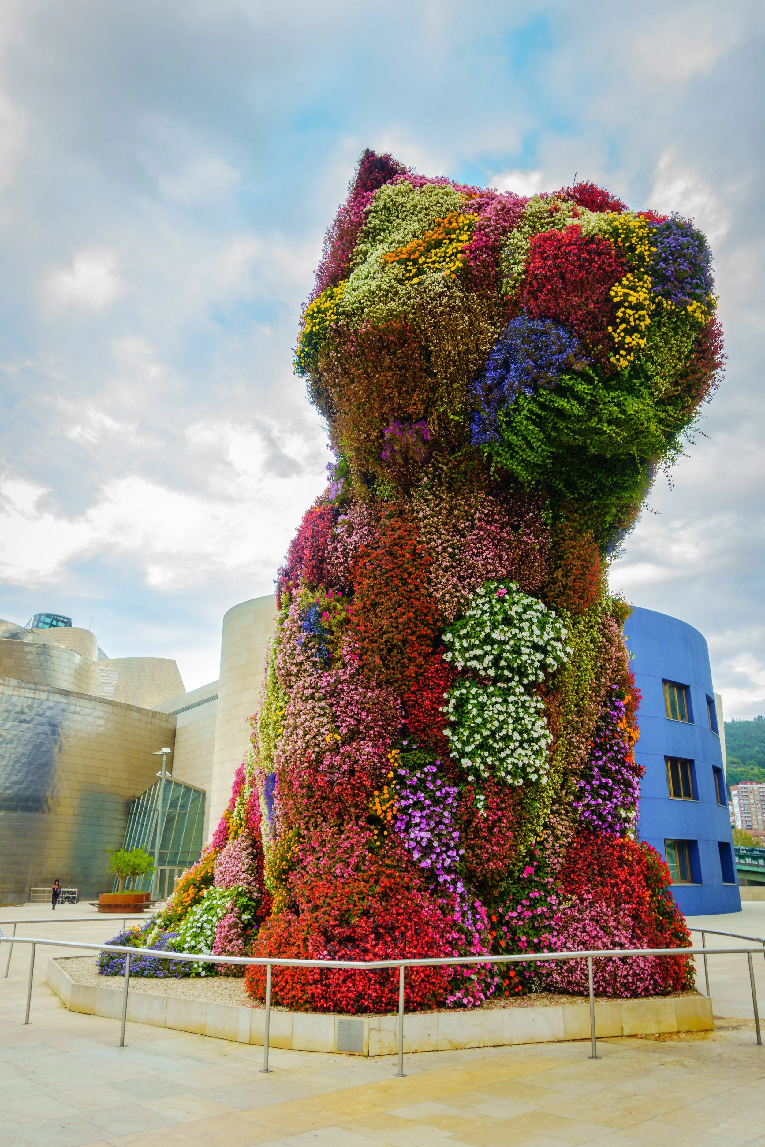 A large, flower-covered bear sculpture outside a modern building with a blue circular section. The sculpture is made entirely of colorful flowers and is set against a partly cloudy sky.