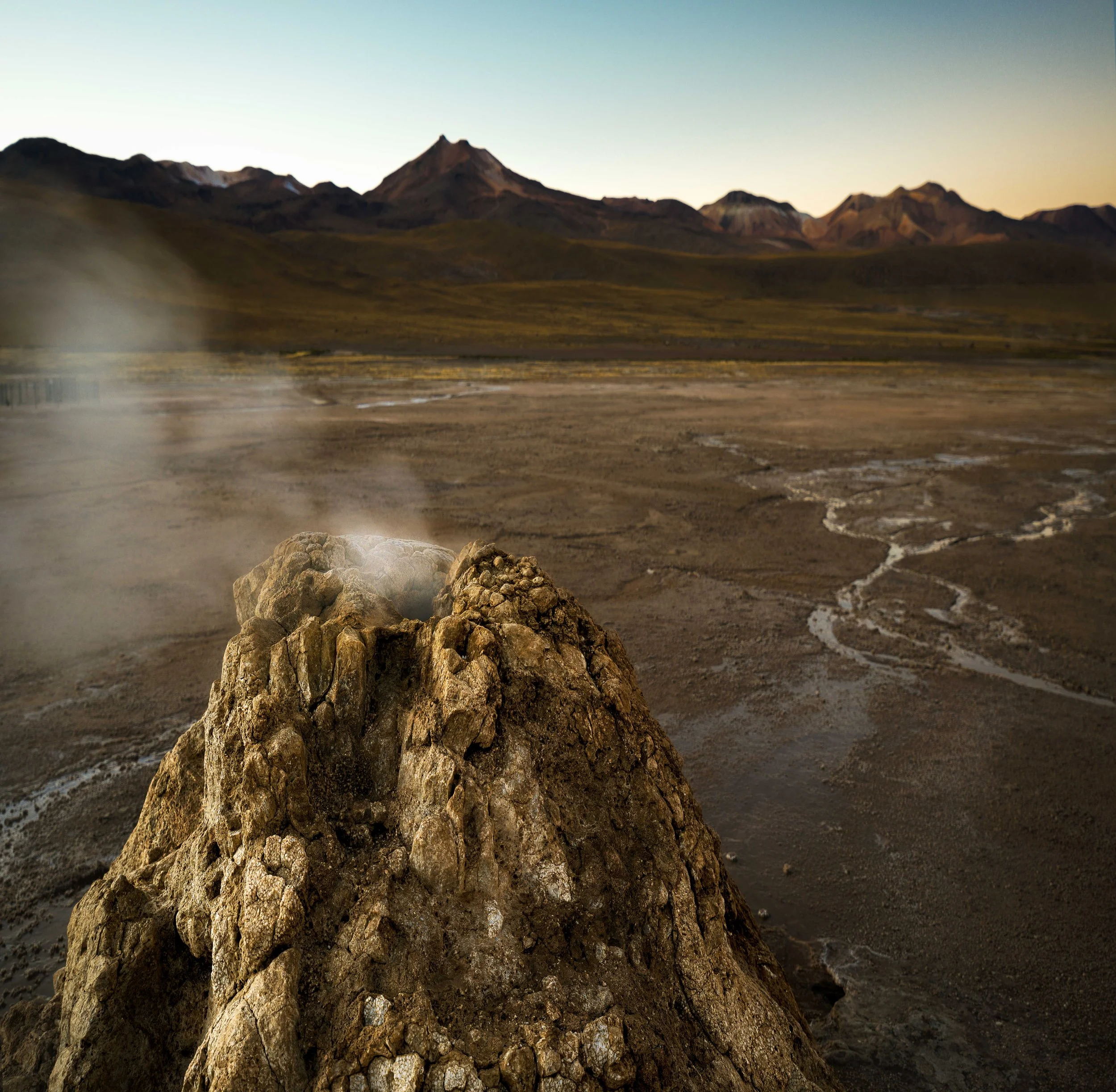 A geothermal steam vent on a barren desert landscape with mountains in the background.
